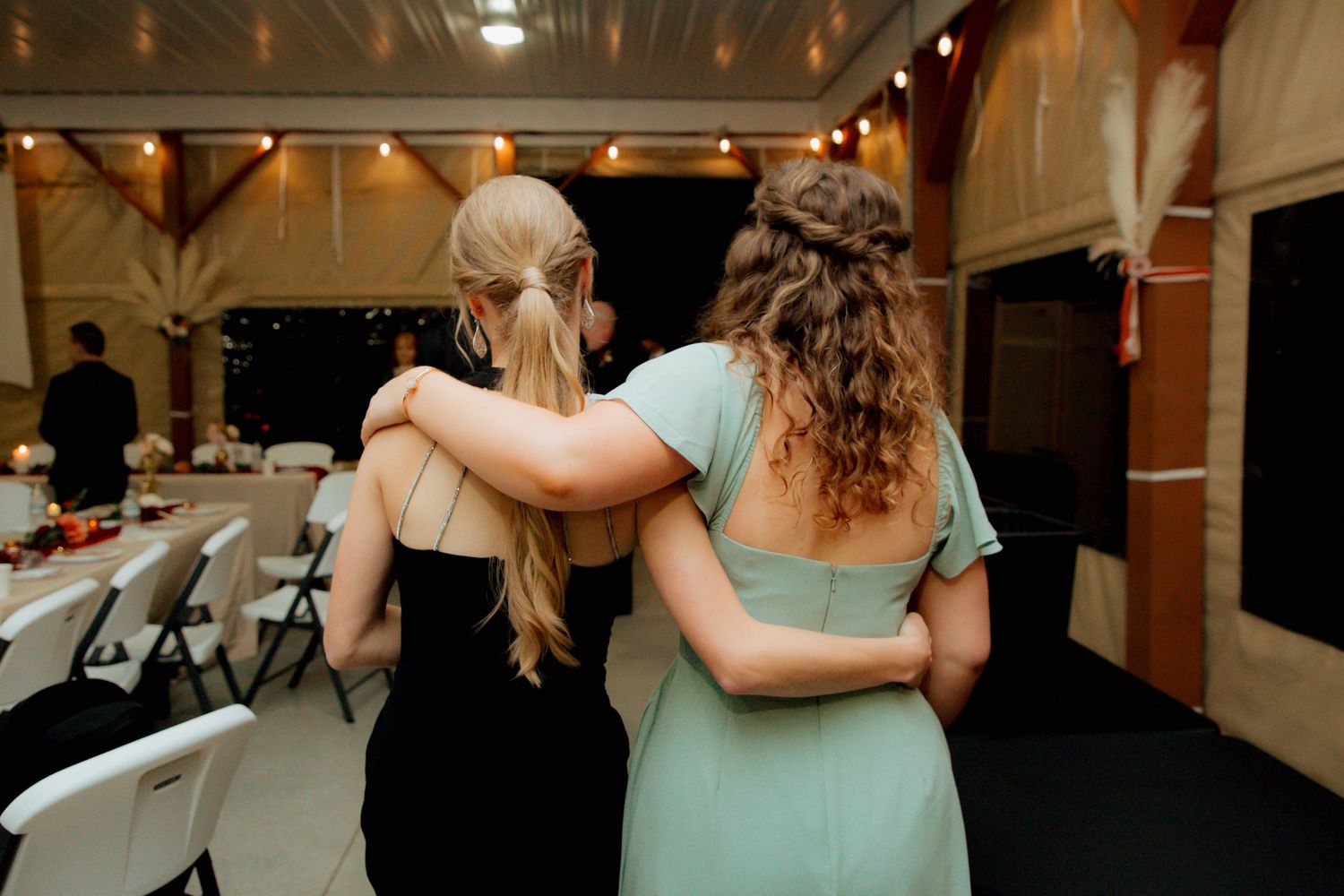 Two women in black and mint green dresses walk arm in arm through barn reception.