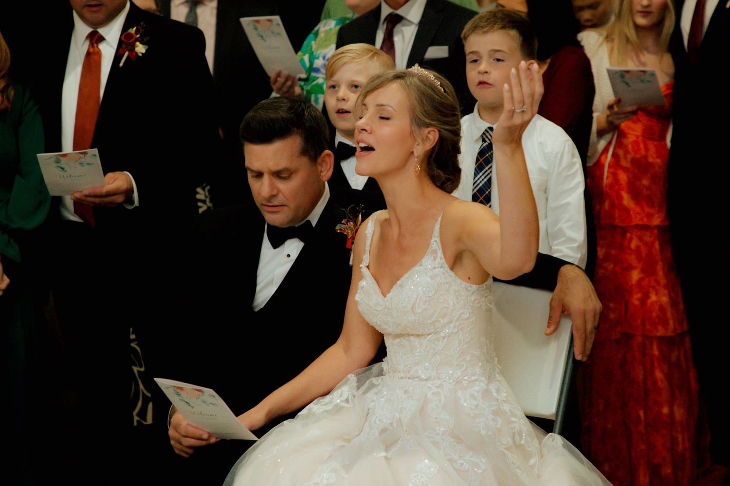 Bride in white ball gown singing joyfully during wedding ceremony with guests.