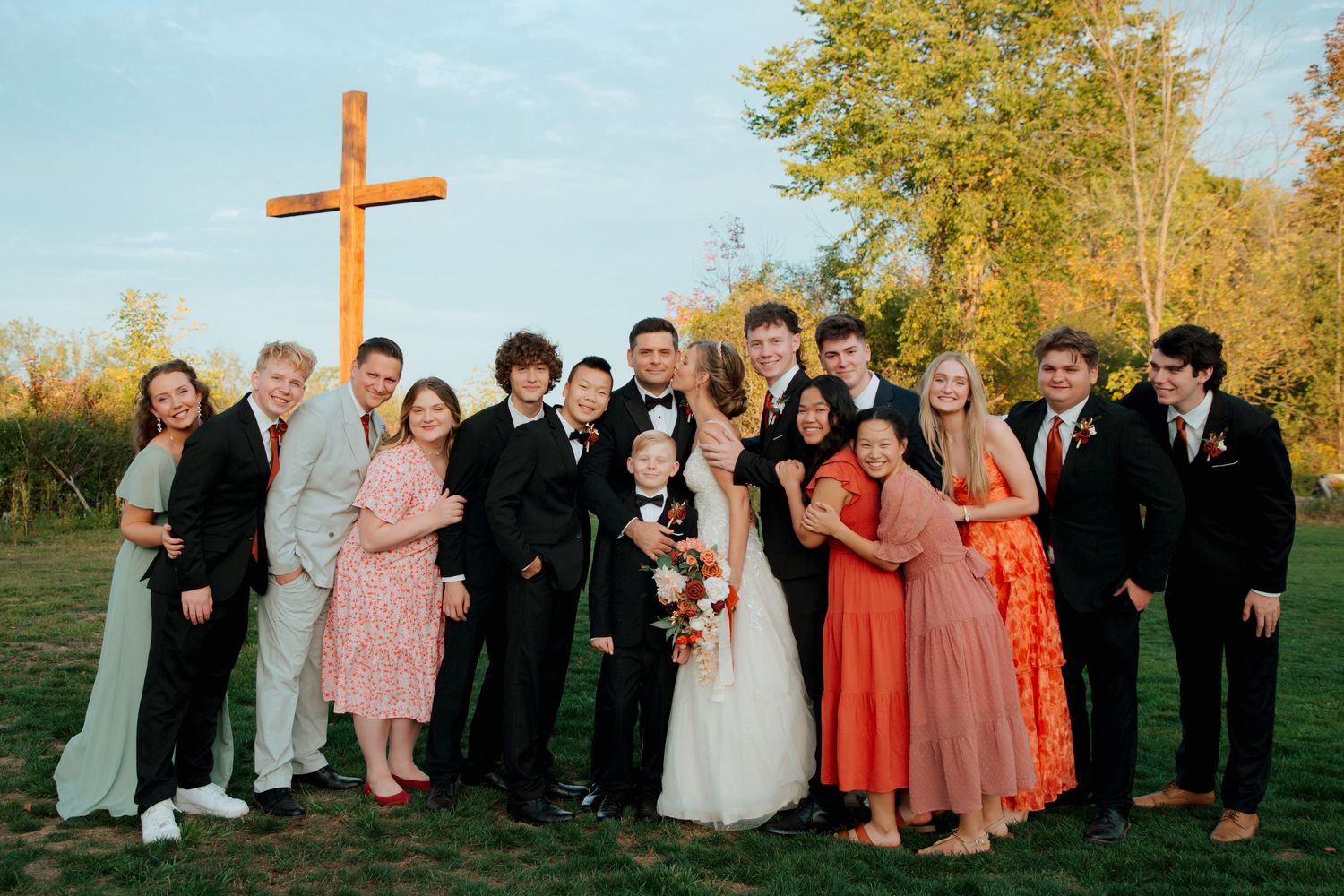 Bride and groom surrounded by teenage wedding party guests near a wooden cross outdoors.