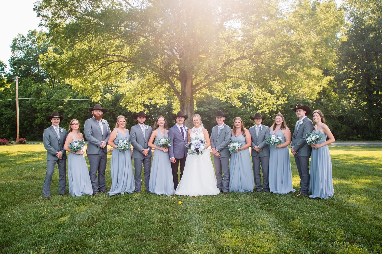 Large wedding party poses in gray suits and dusty blue gowns on green lawn beneath a grand tree.