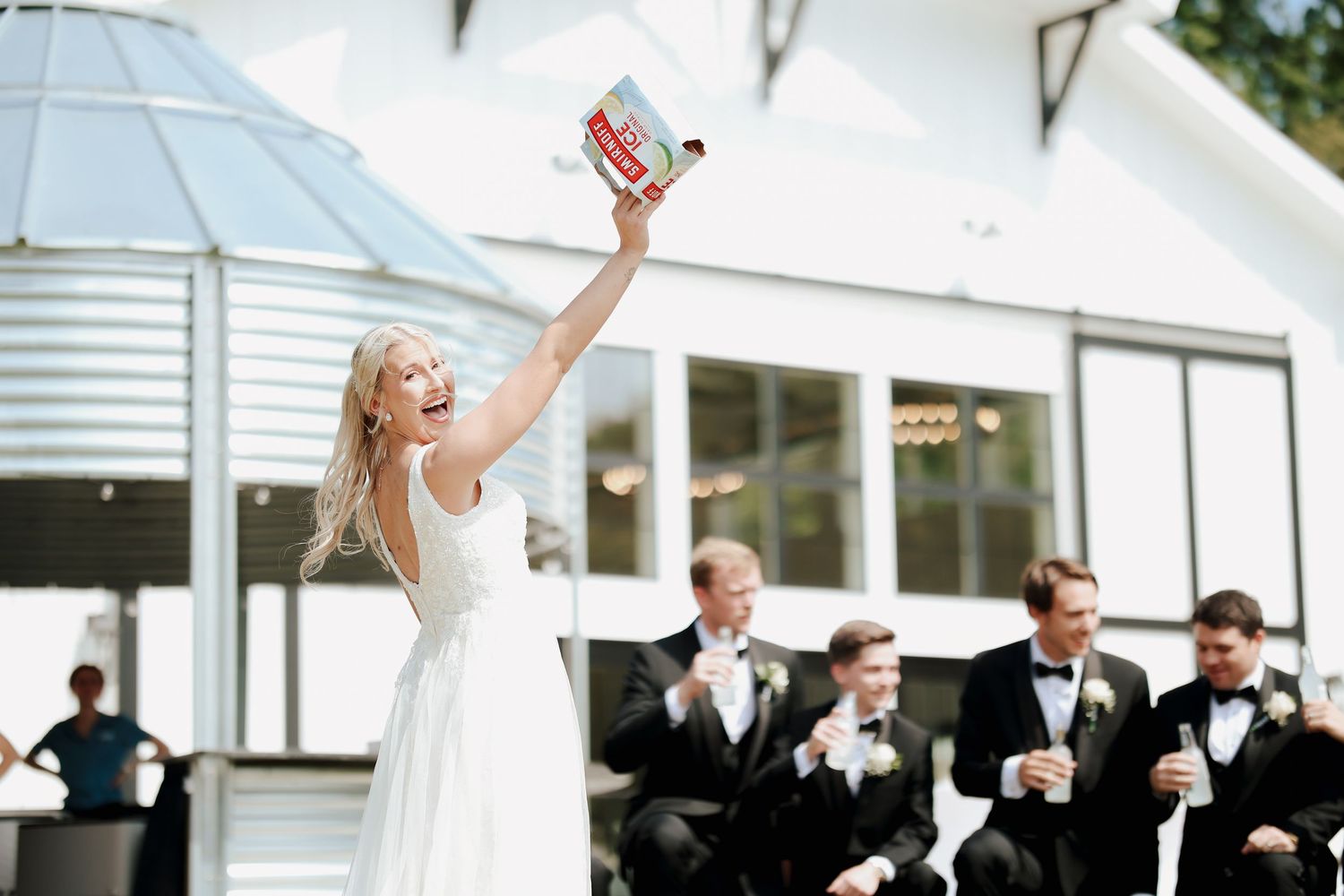 Laughing bride raises a box of Cheez-Its triumphantly while groomsmen celebrate at outdoor wedding venue.