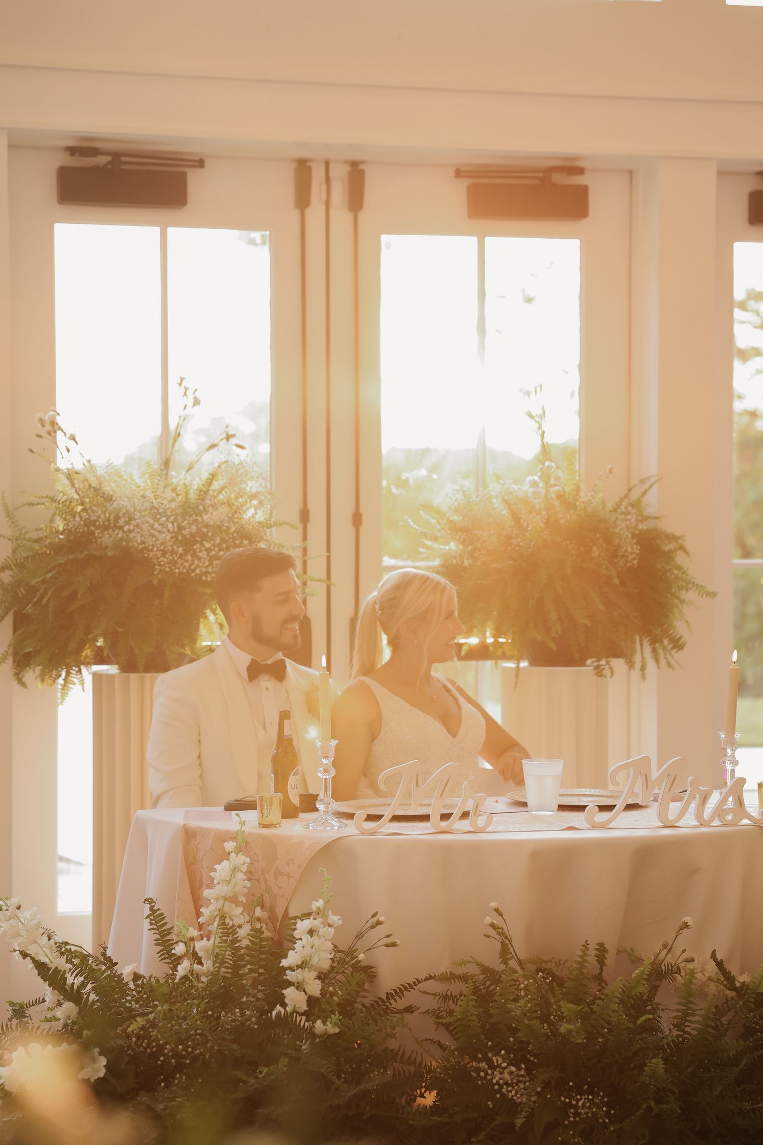 Bride and groom seated at sweetheart table bathed in golden sunlight at wedding reception.