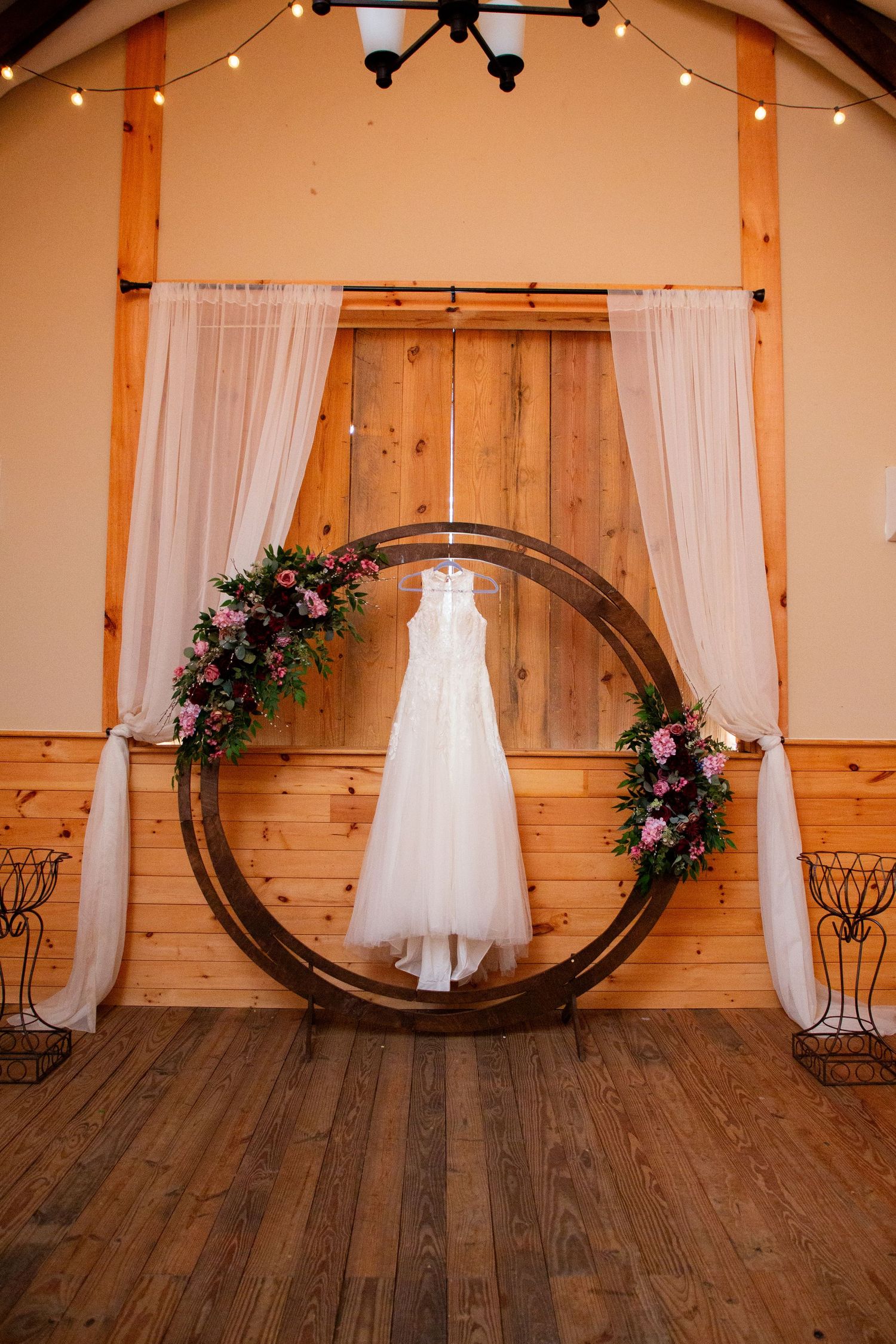 White wedding dress hanging in circular wooden arch with pink floral arrangements in rustic barn venue.