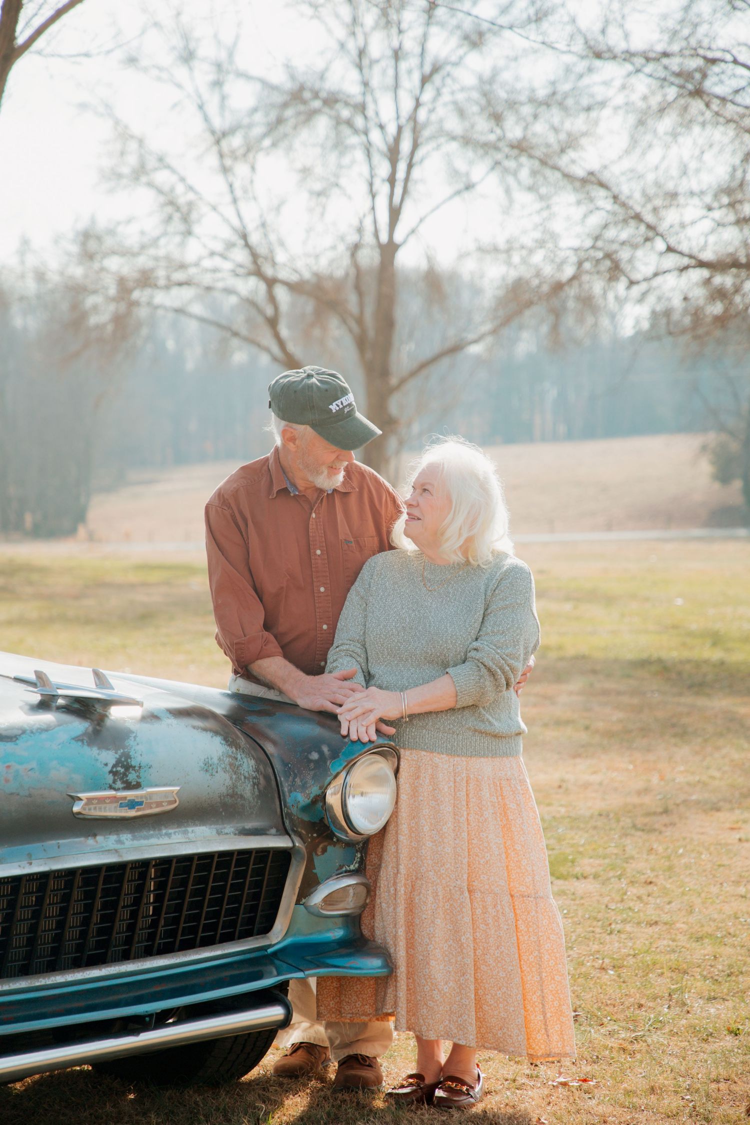 Elderly couple sharing a tender moment leaning against a vintage teal classic car in a sunny park.