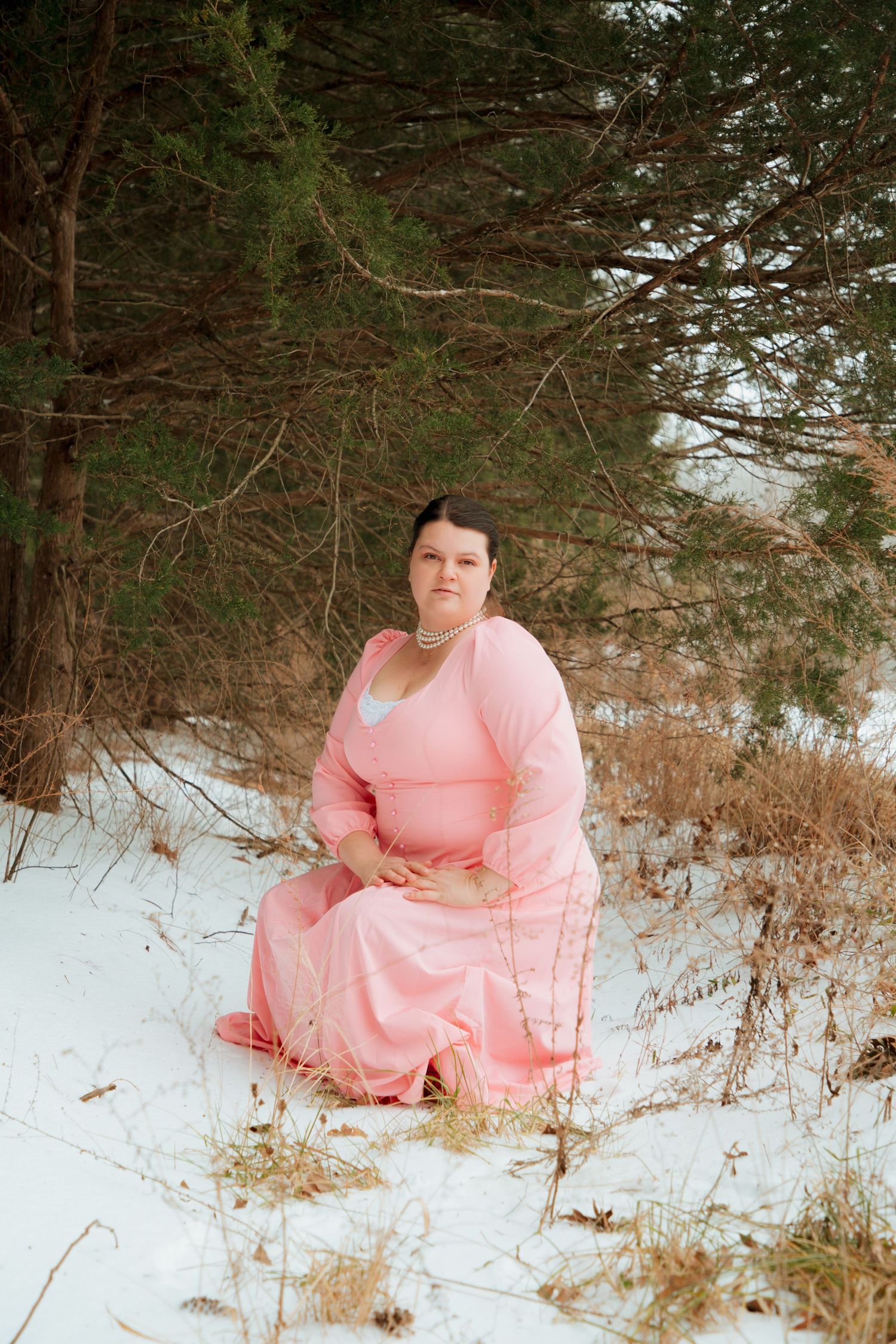 Plus-size woman in long pink dress sitting gracefully in a snowy pine forest setting.