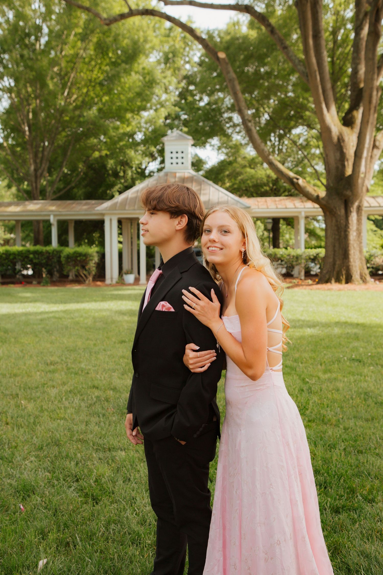 Teen prom couple posing back-to-back on green lawn with white gazebo in background.