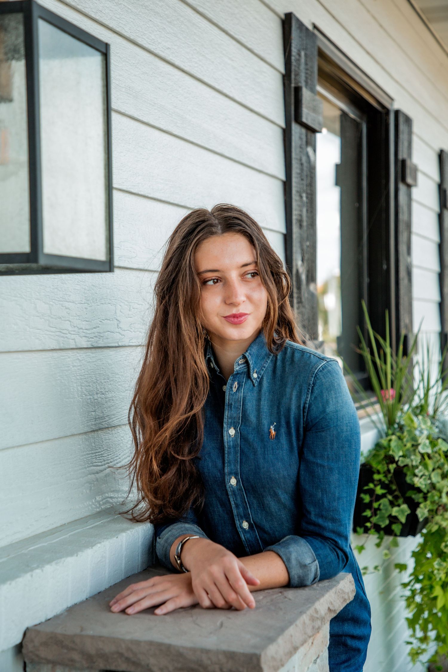 Young woman with long brown hair in denim shirt leaning on stone ledge outside white house.