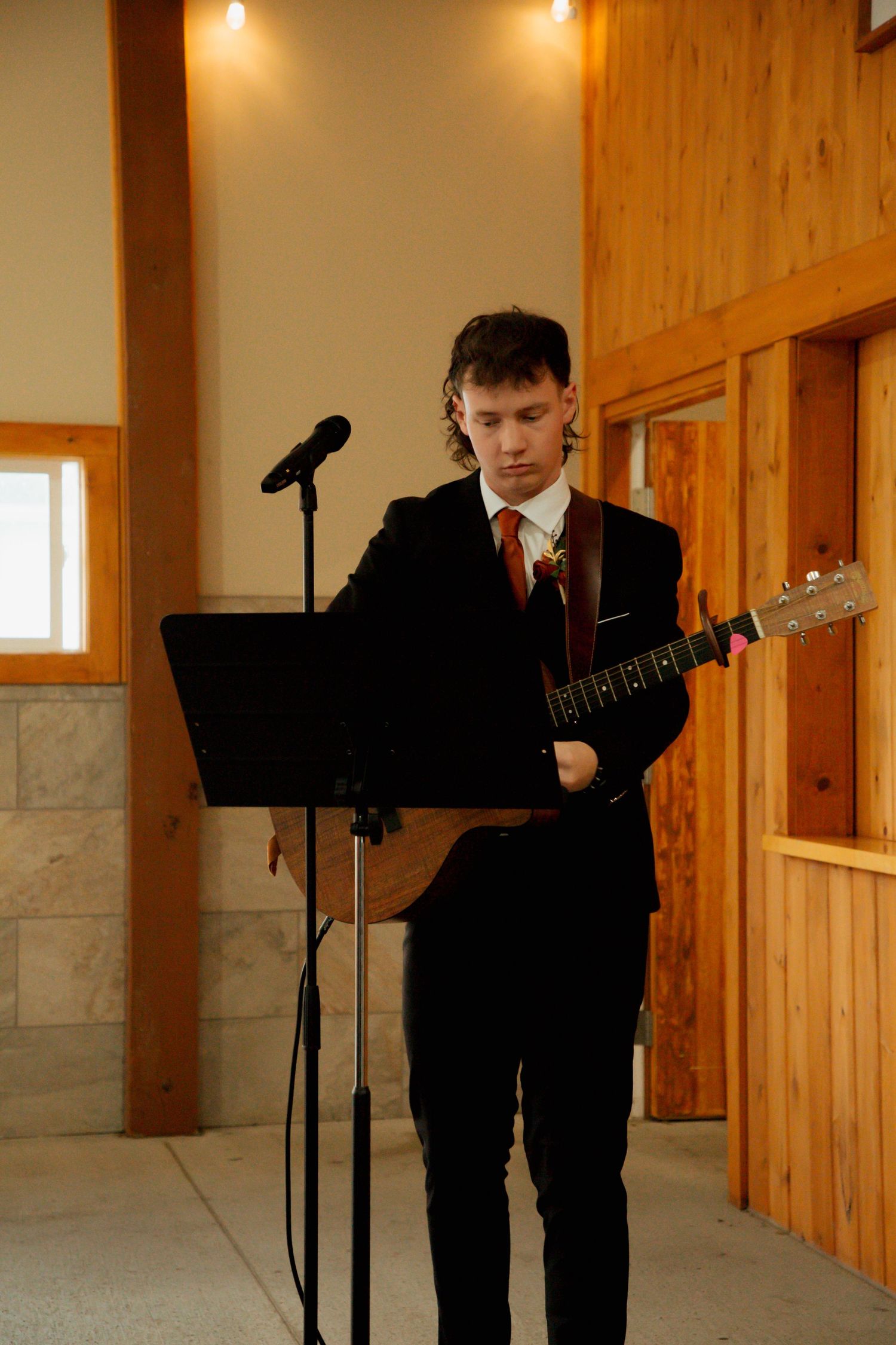 A young man in a black suit plays acoustic guitar at a wedding ceremony microphone stand.