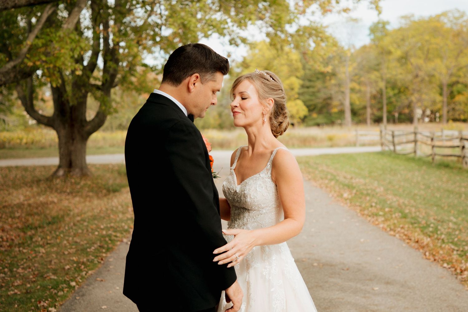 Bride in lace gown and groom in tuxedo sharing intimate moment on fall wedding path.