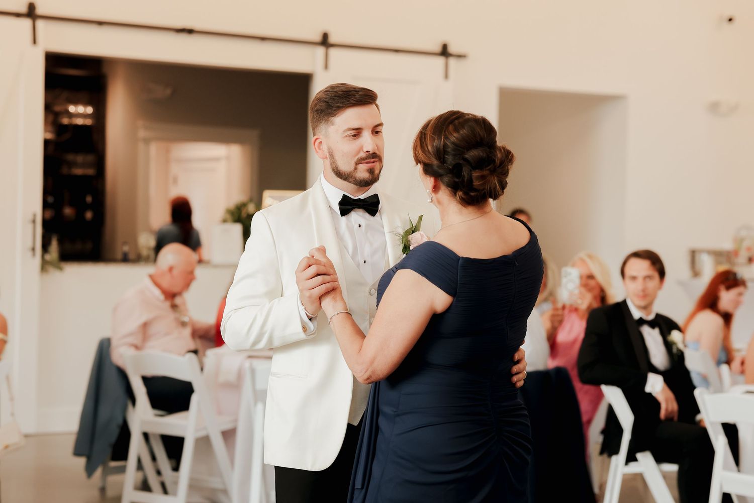 Groom in white tuxedo dances with mother in navy gown at formal wedding reception.