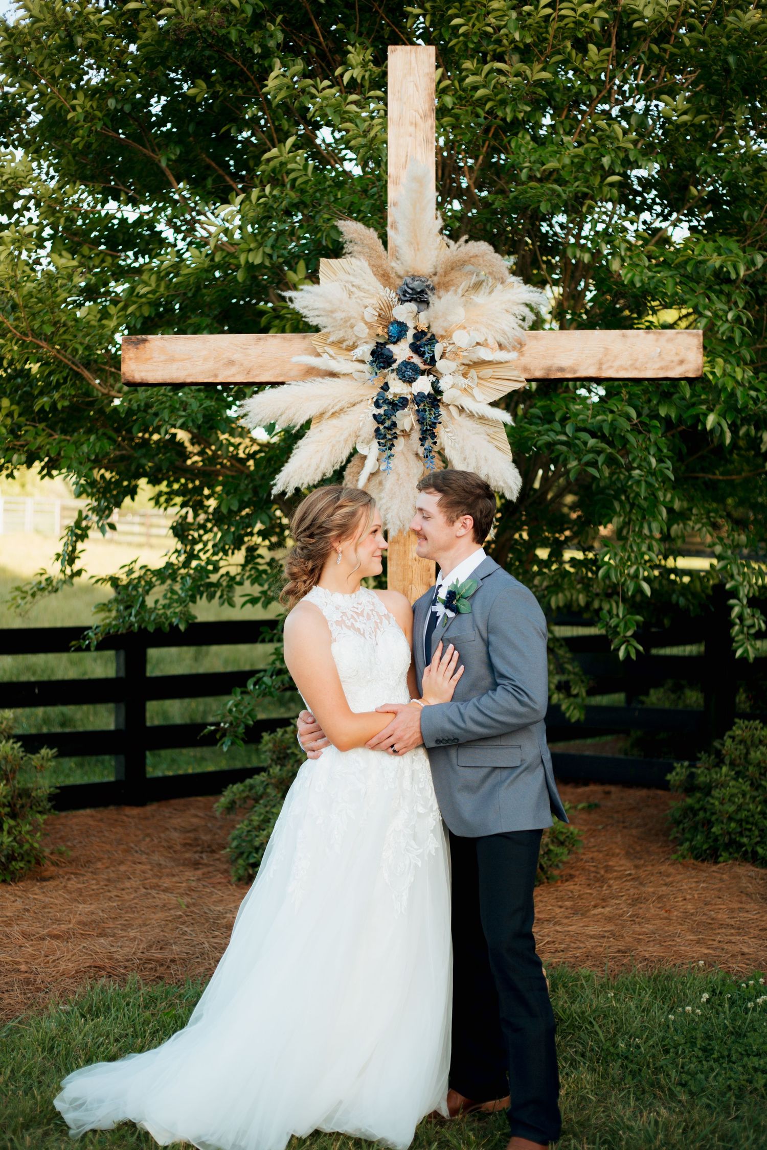 Bride and groom share a romantic moment in front of a rustic wooden cross decorated with pampas grass at outdoor wedding.