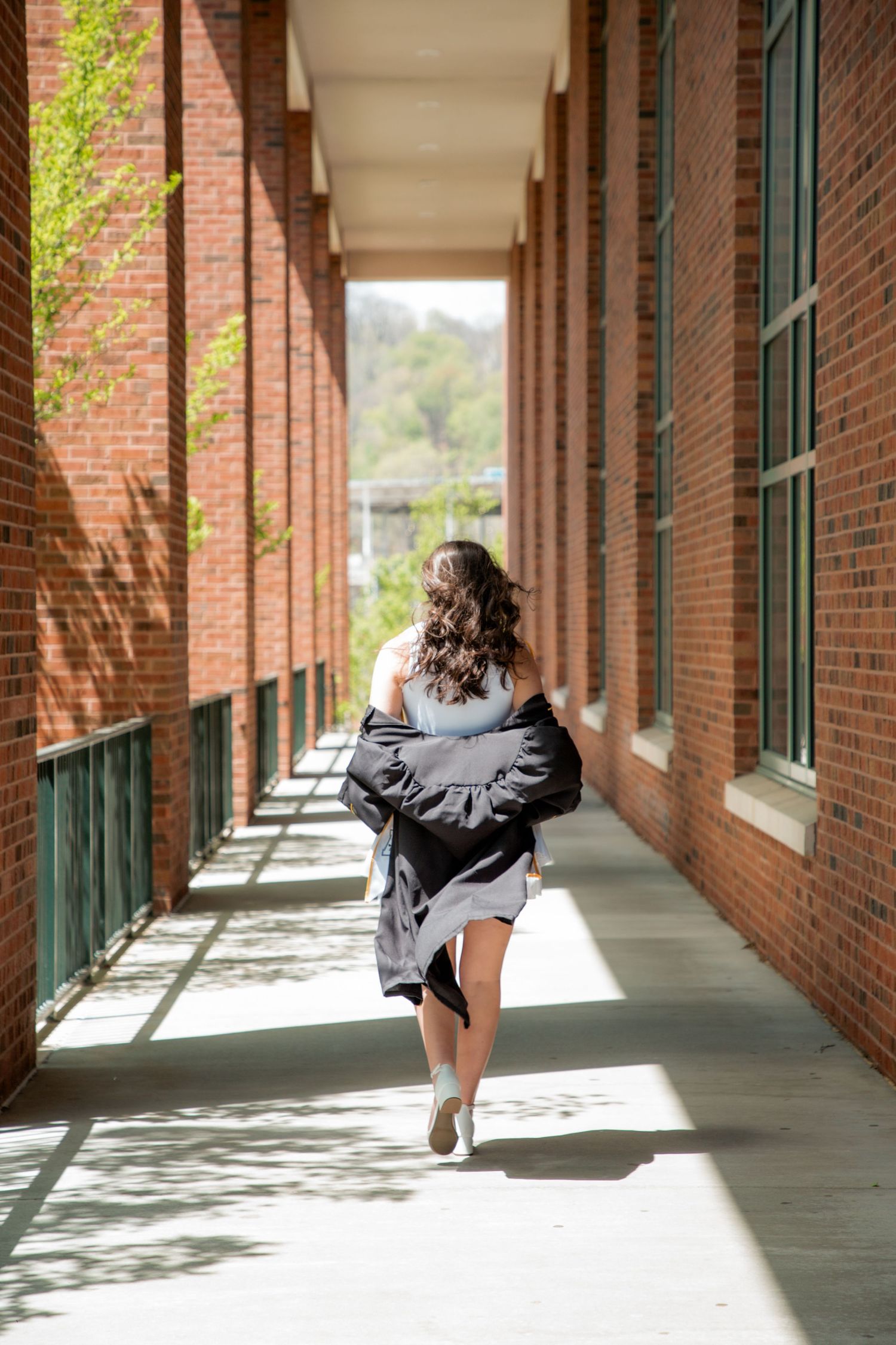 Graduate walking away in black gown and white dress through sunny brick corridor at Appalachian State.