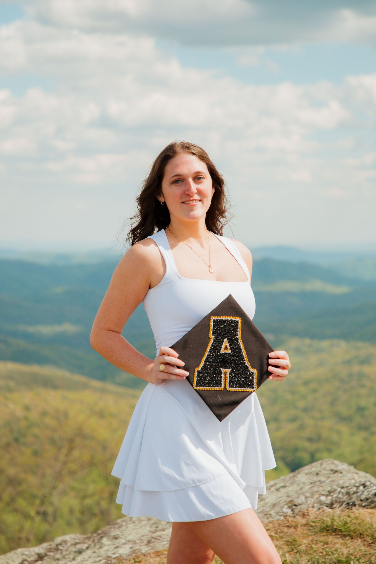 Smiling graduate in white dress holding Appalachian State graduation cap on scenic mountain overlook.