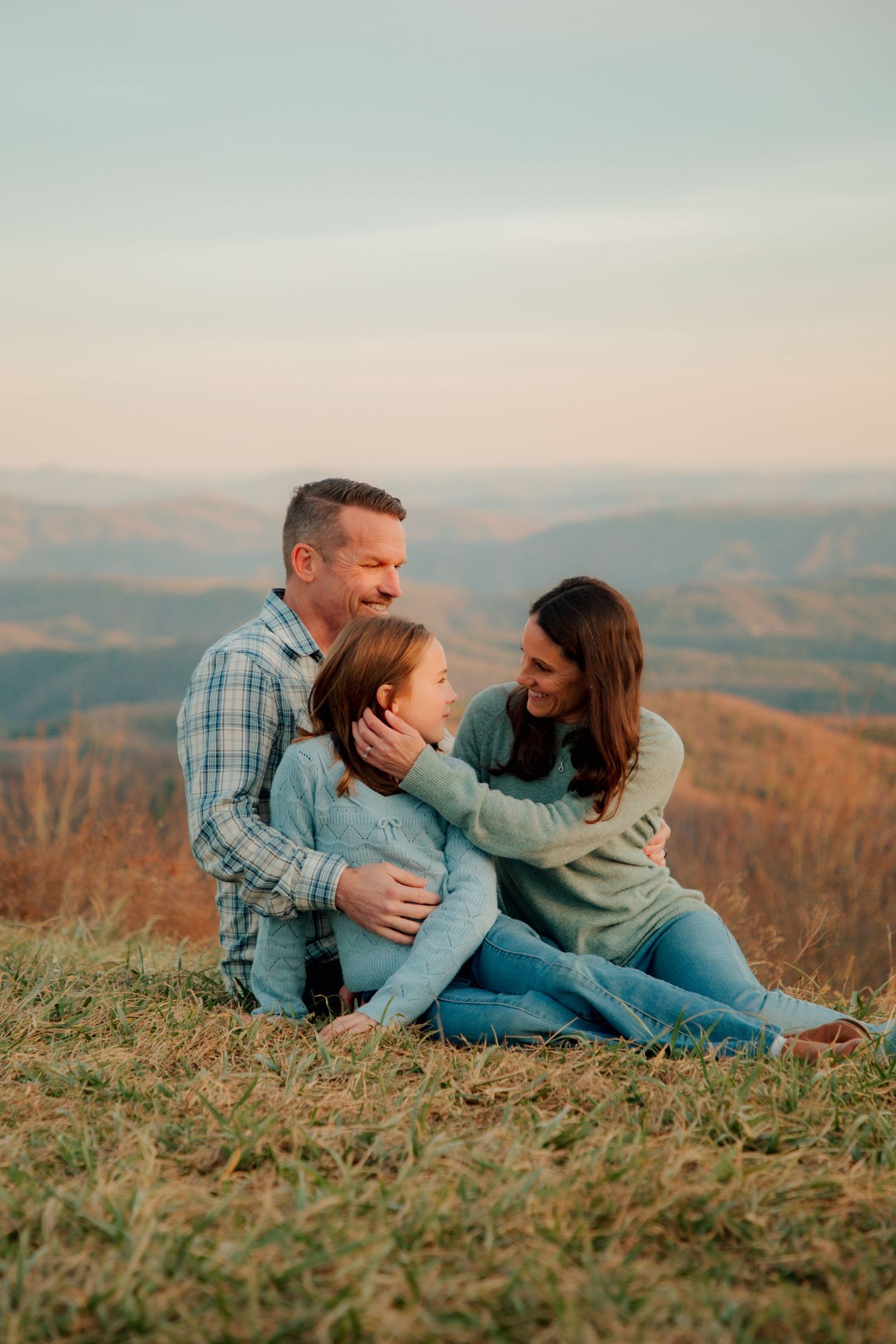 A cheerful family of three sits on a hillside, sharing a joyful moment with rolling mountains in the background.
