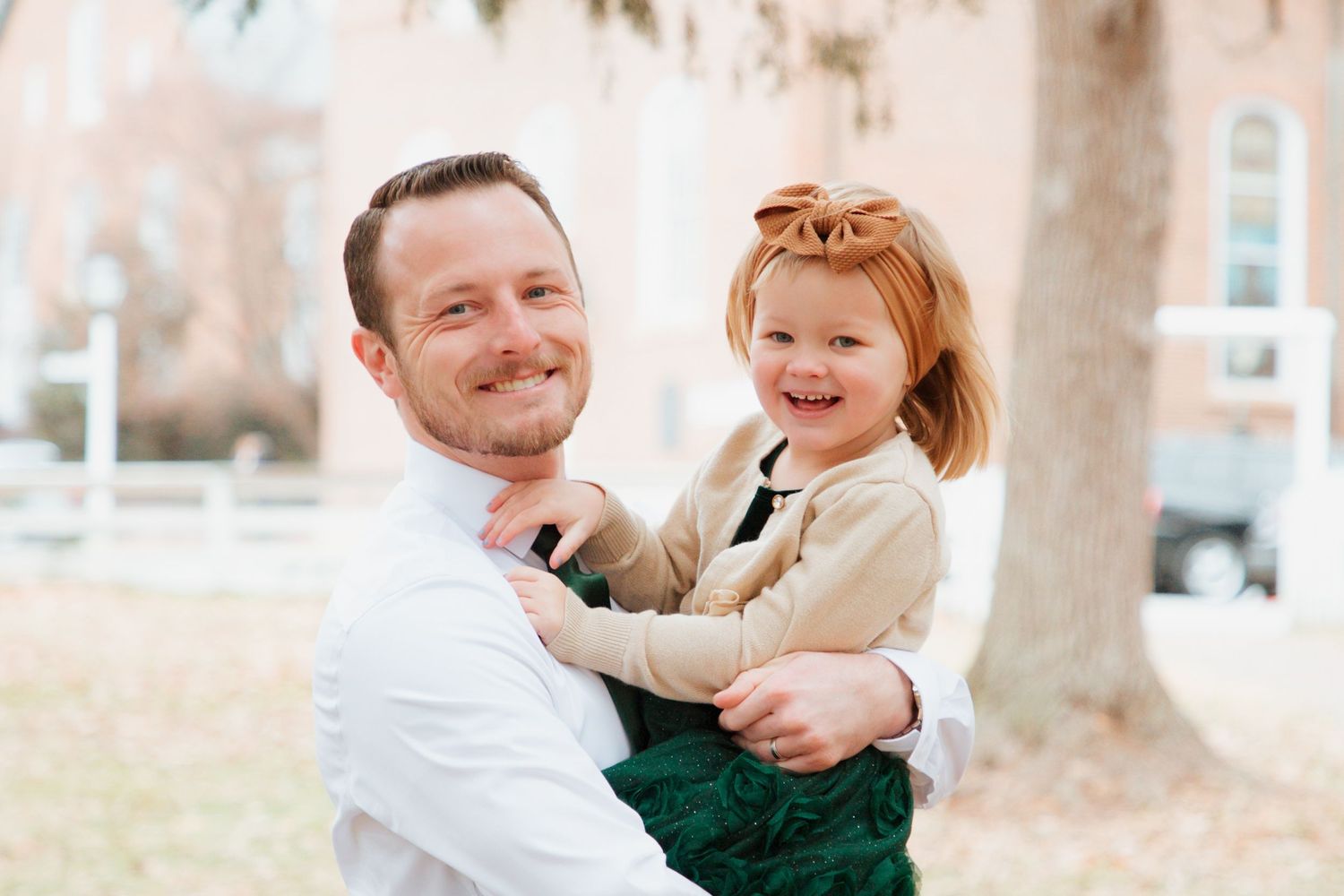 Smiling father holding happy red-haired toddler daughter in green dress during outdoor family photos.