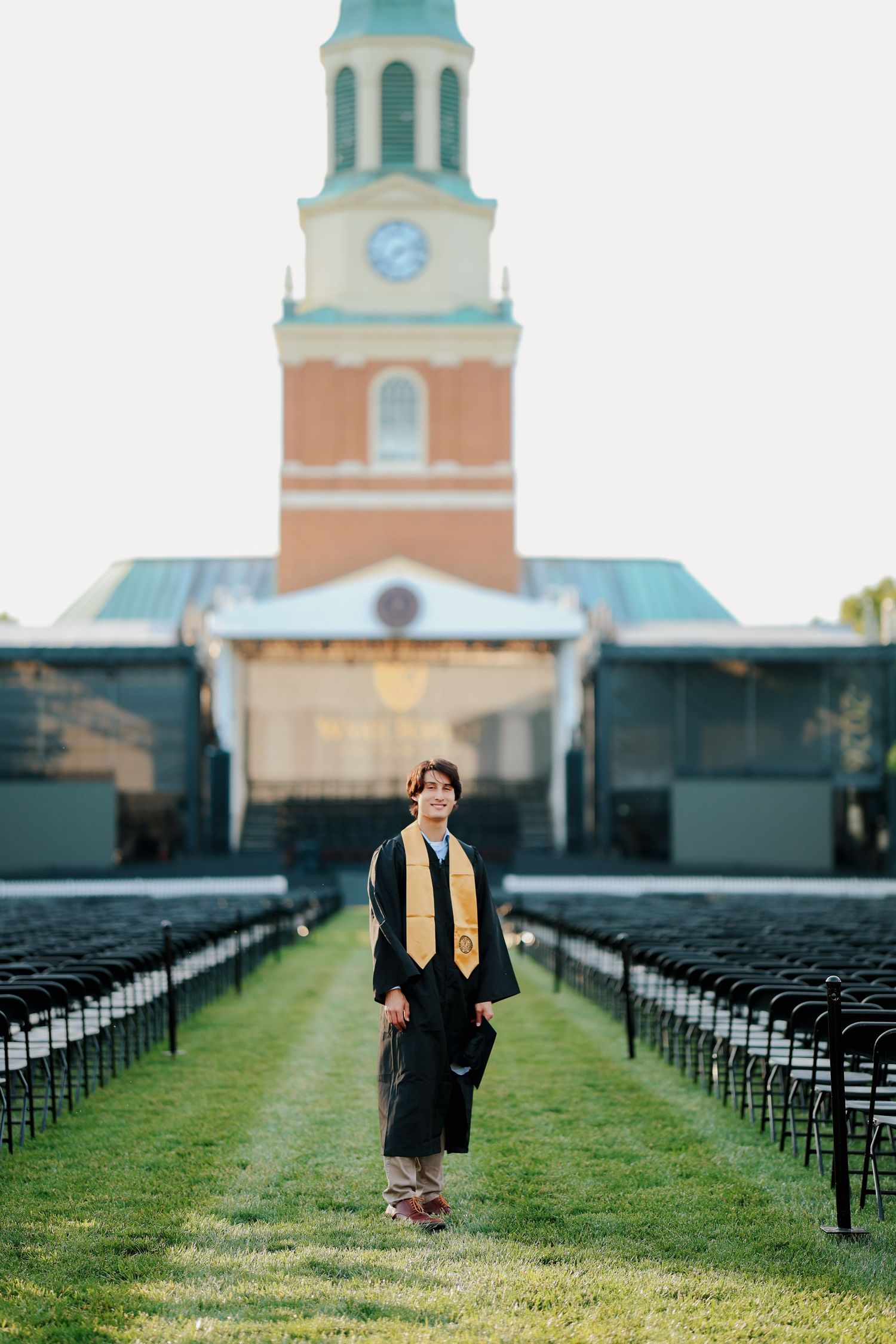Graduate in black cap and gown with gold stole walks between rows of chairs at ceremony venue.