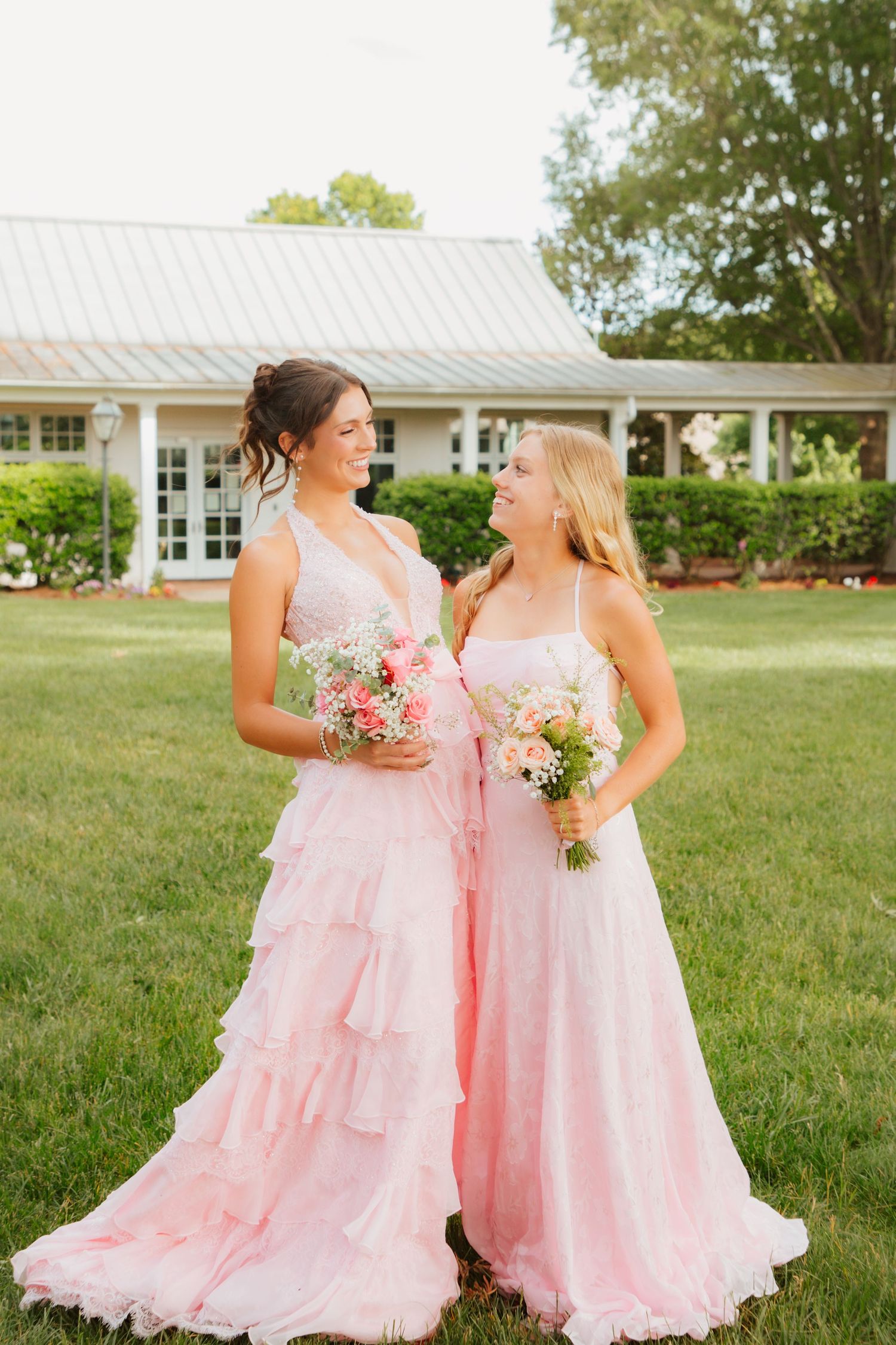 Two young women in matching pink formal gowns holding bouquets, smiling at each other on green lawn.