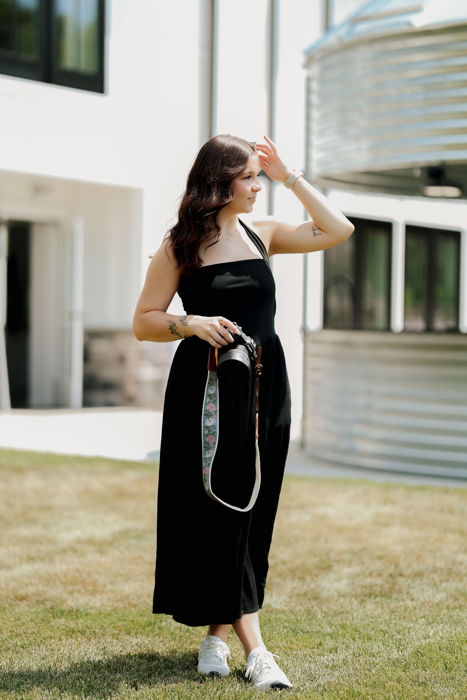 Young woman in black midi dress holding camera with floral strap outdoors in bright sunlight.