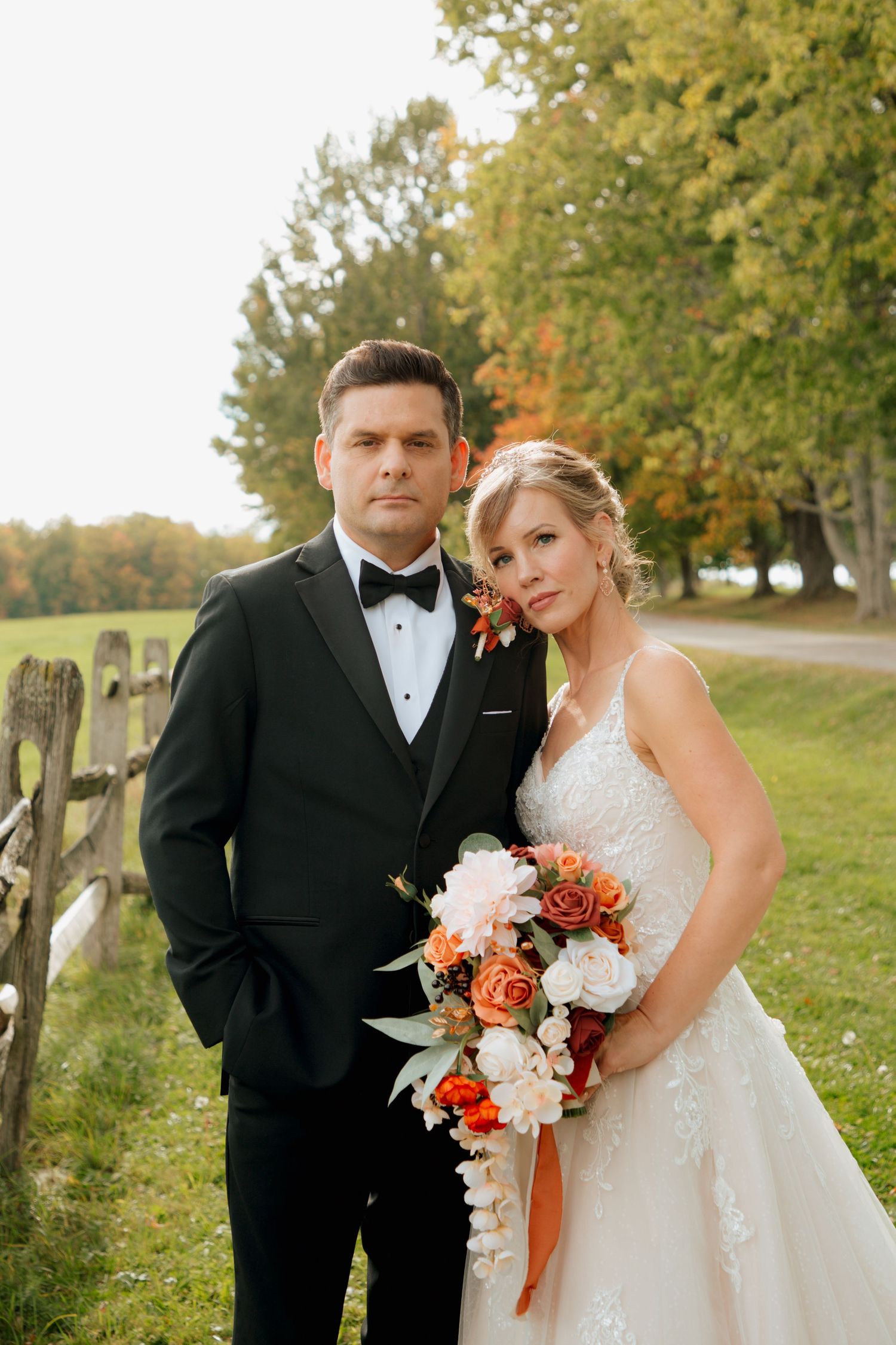 Elegant couple in wedding attire posing closely by a wooden fence with fall foliage backdrop.