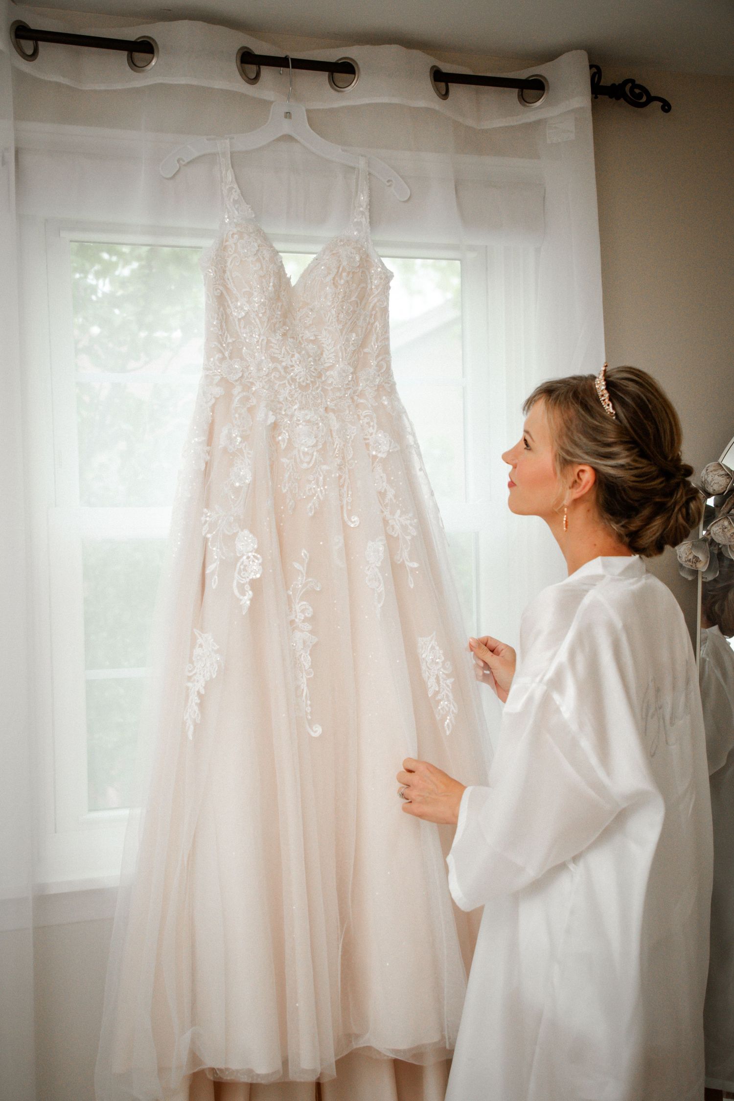 Bride in white robe admiring her lace blush wedding dress hanging by window.