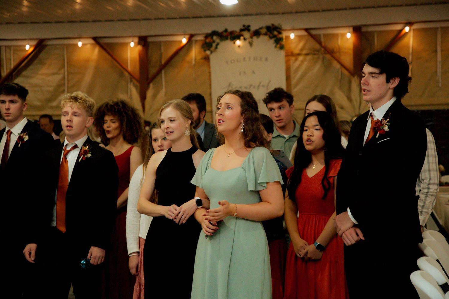 Young wedding guests in formal attire singing together at rustic barn reception.