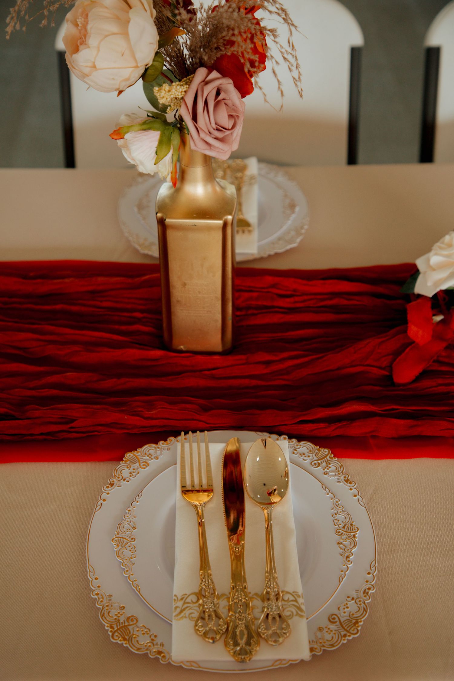 Formal wedding place setting with gold cutlery, ornate white plate, and gold vase with pink and cream flowers on red runner.