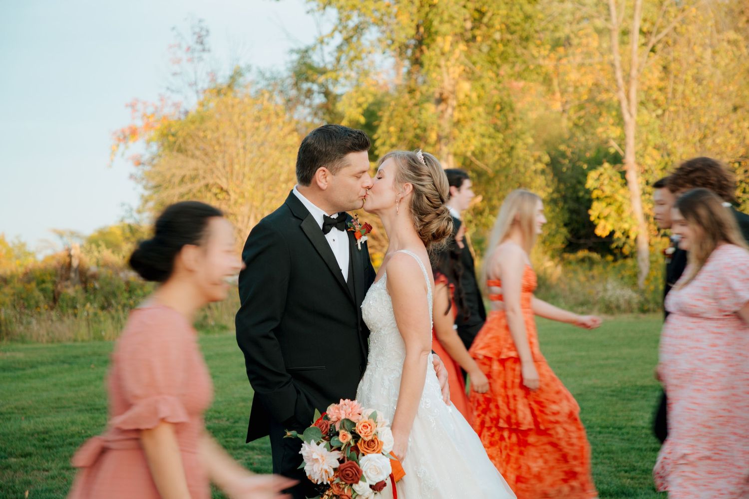 Bride and groom sharing a kiss outdoors while wedding guests in colorful dresses walk by.