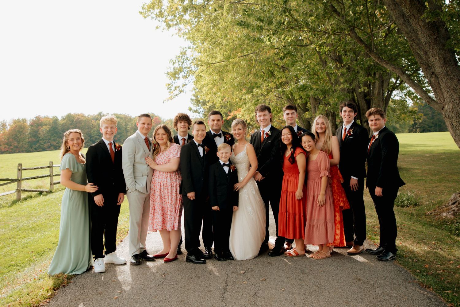 Large wedding party group photo on a tree-lined path featuring bride, groom, and family members in fall attire.