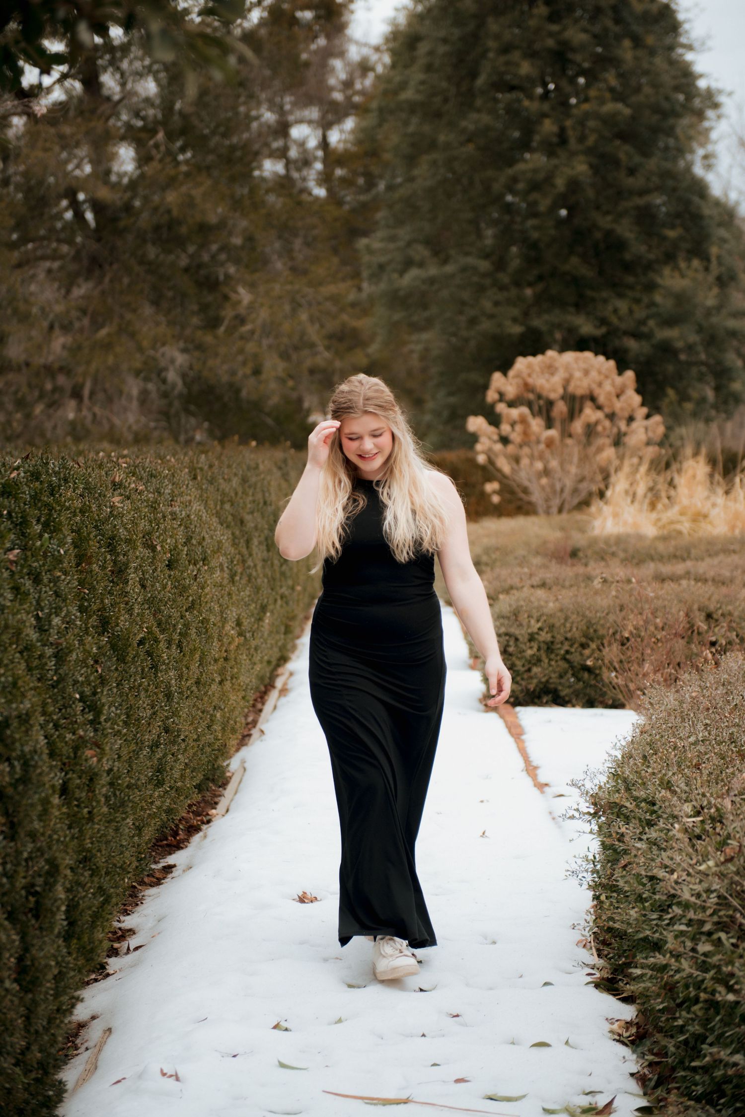 Young woman in a black maxi dress walking on a snow-dusted garden path lined with green hedges.