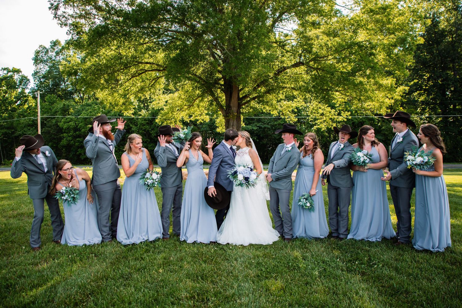 Wedding party laughs and kisses in a lively outdoor group photo beneath a large tree in golden sunlight.