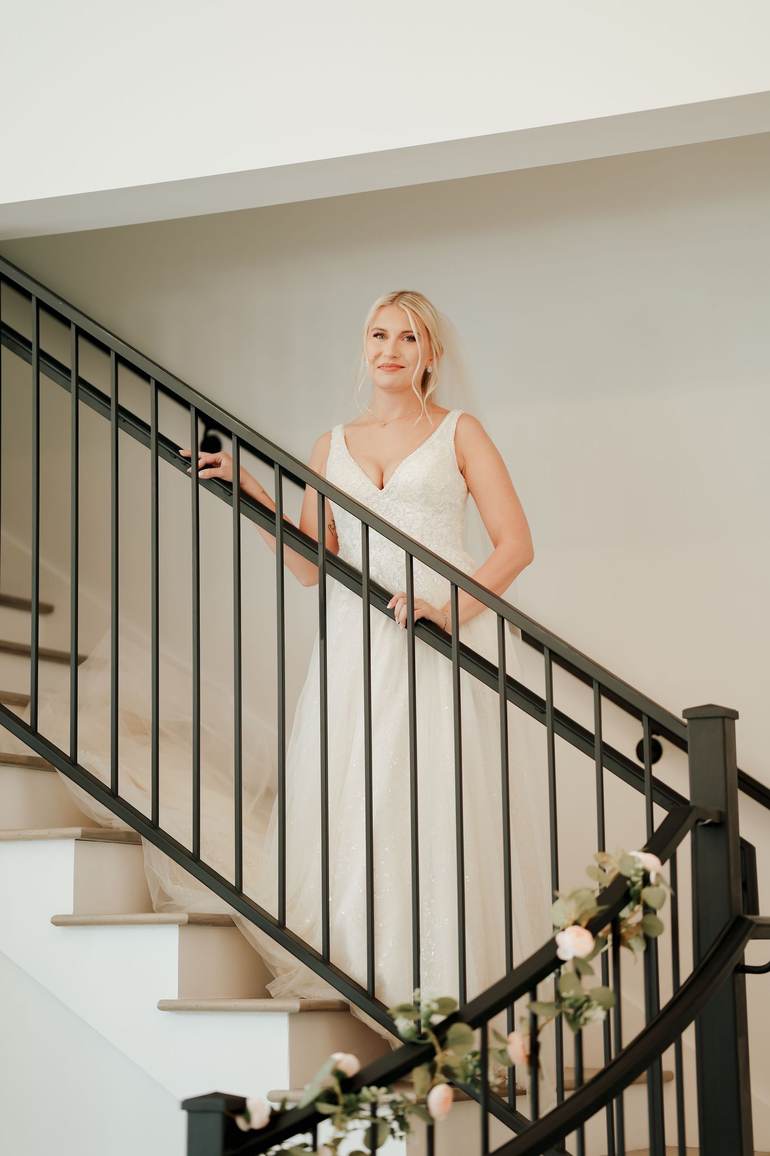 Beautiful blonde bride in fitted white gown posing gracefully on staircase decorated with floral garland.