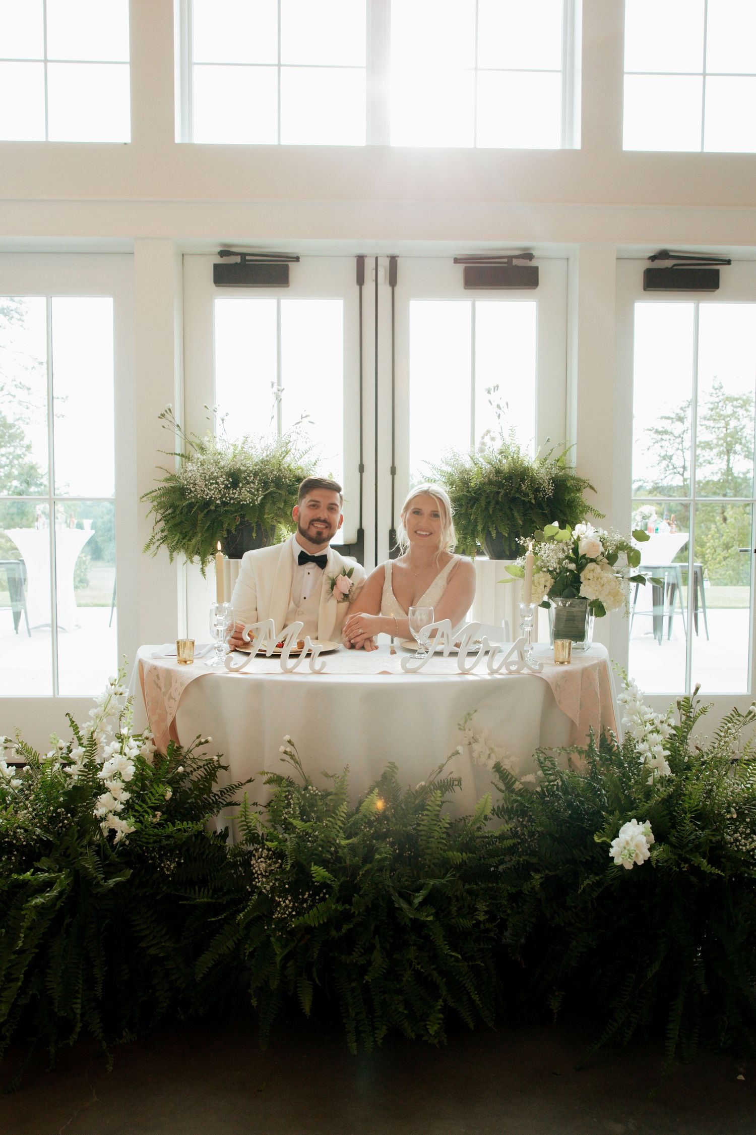 Smiling bride and groom at sweetheart table surrounded by lush greenery and white flowers.