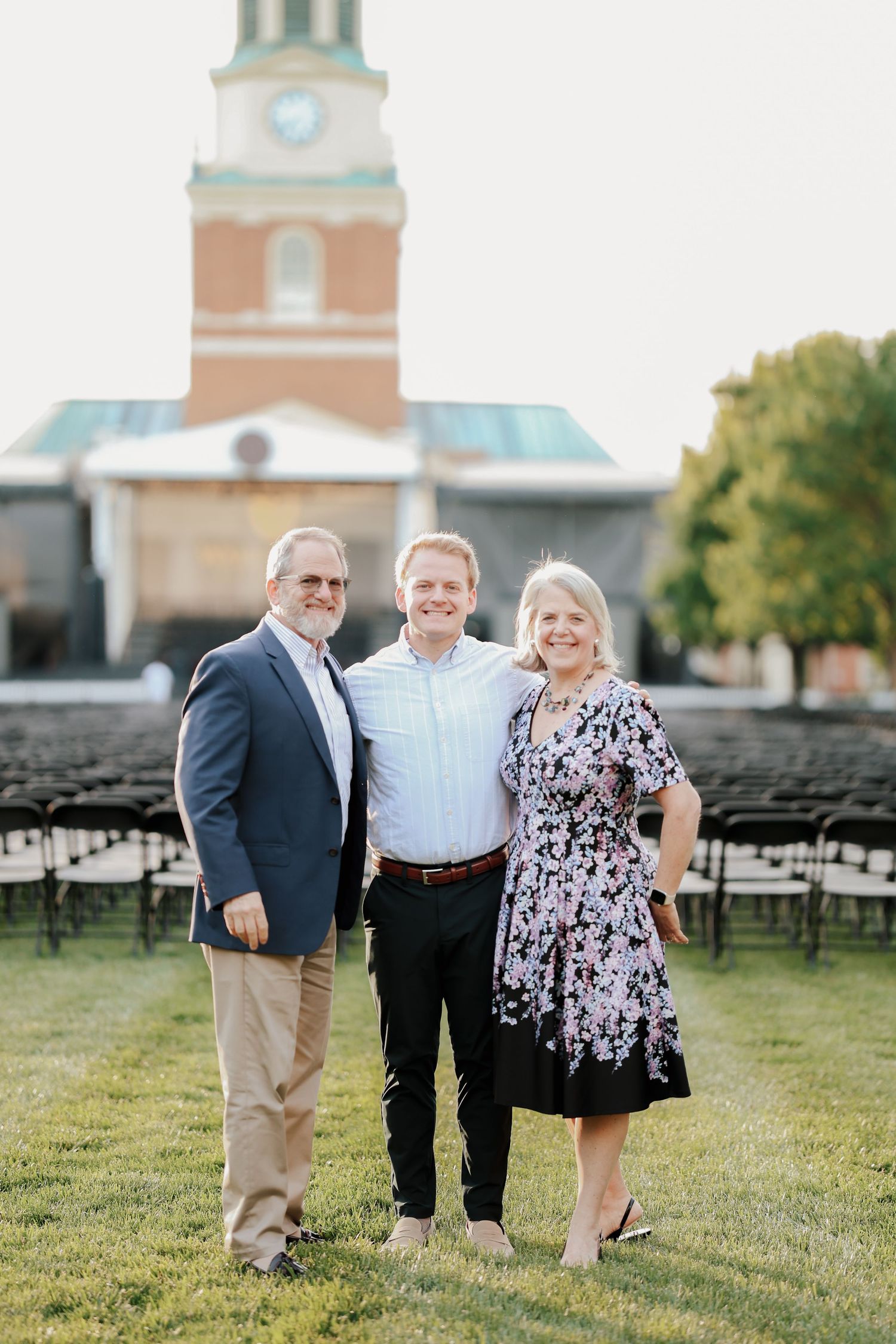 Family of three smiling together on green lawn with brick clock tower building in background.