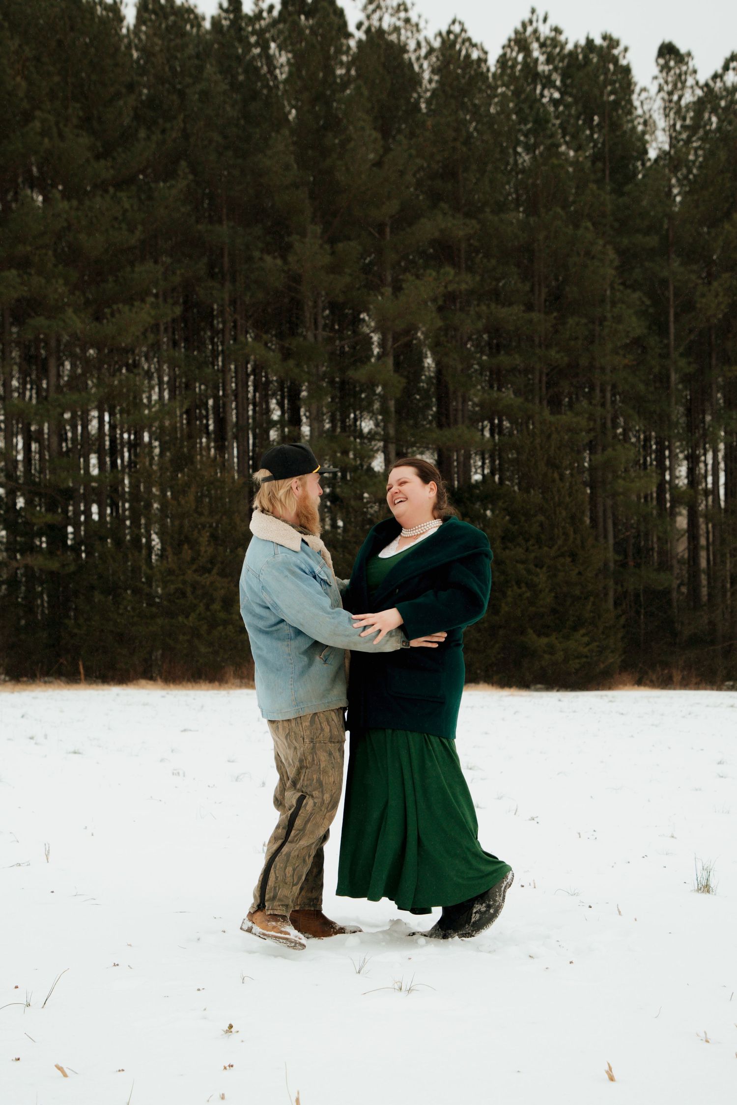Laughing couple holding hands in snowy field, surrounded by tall evergreen trees in winter.
