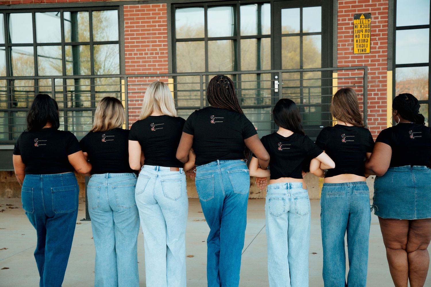 Seven women in matching black t-shirts and jeans standing with backs to camera near large windows.