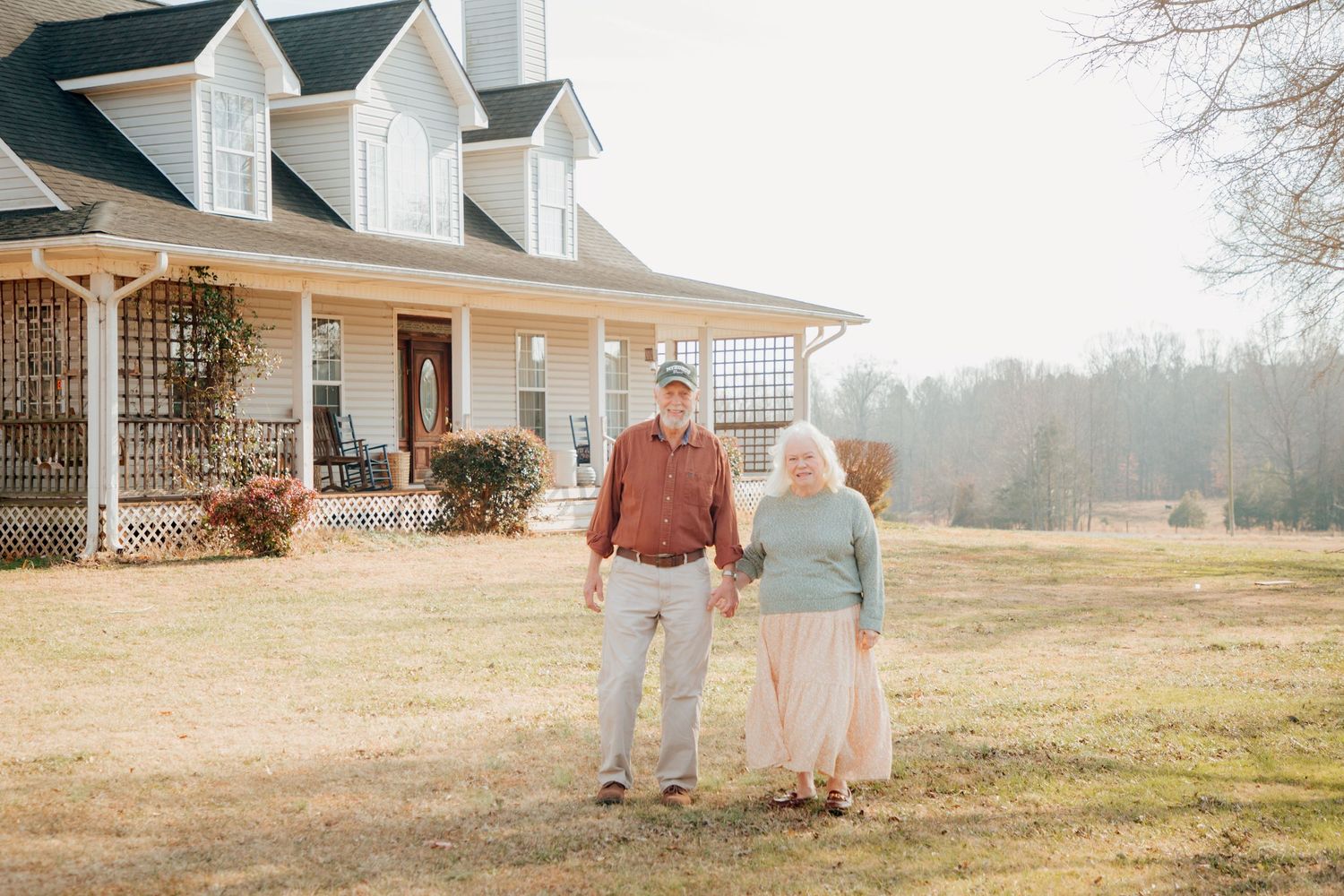 Senior couple holding hands while walking in front of a charming white farmhouse.