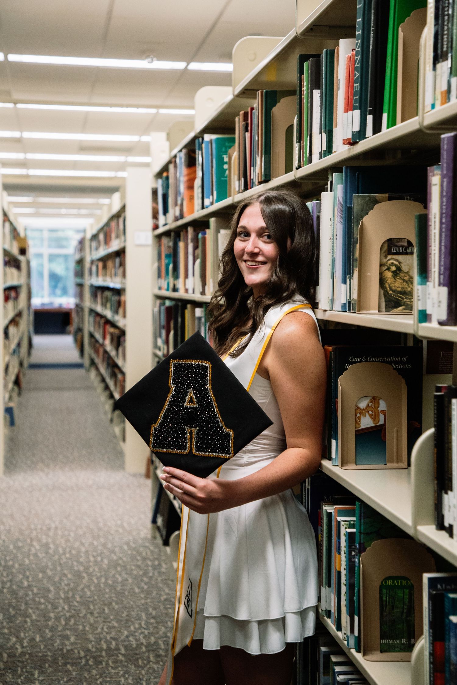 Smiling female graduate in white dress holds black cap with gold A letter in library aisle.