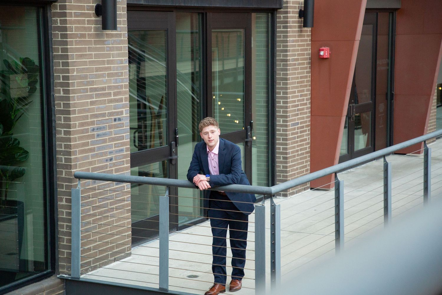 Young man in navy suit leaning on a glass railing outside a modern brick building.