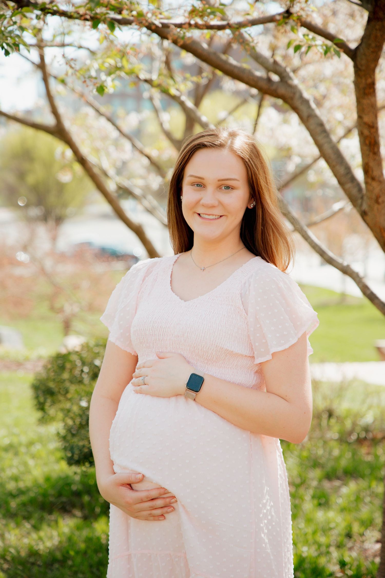 Smiling pregnant woman in pink dress cradling baby bump under flowering tree outdoors.