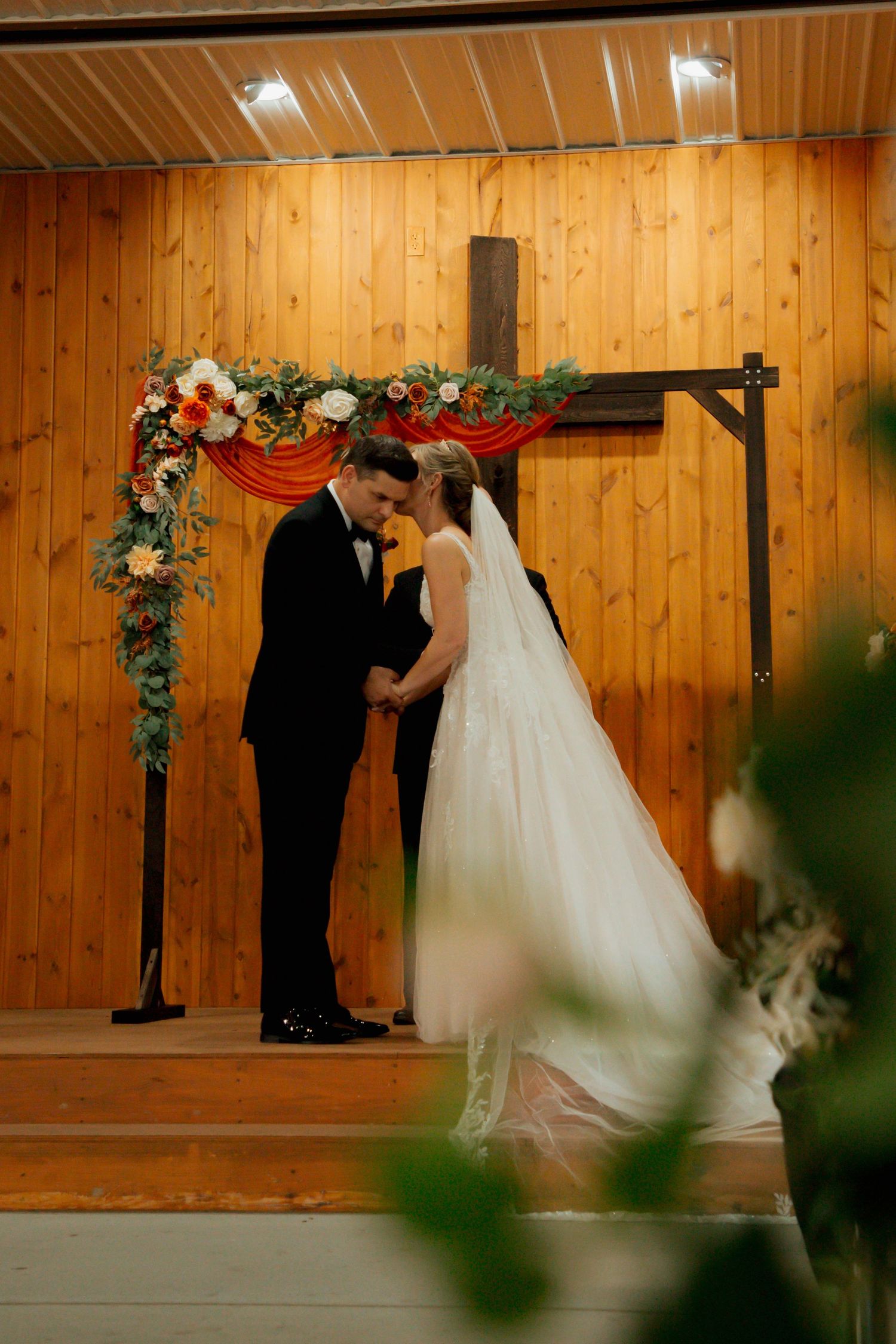 Bride and groom share intimate moment holding hands beneath decorated wooden cross wedding arch.