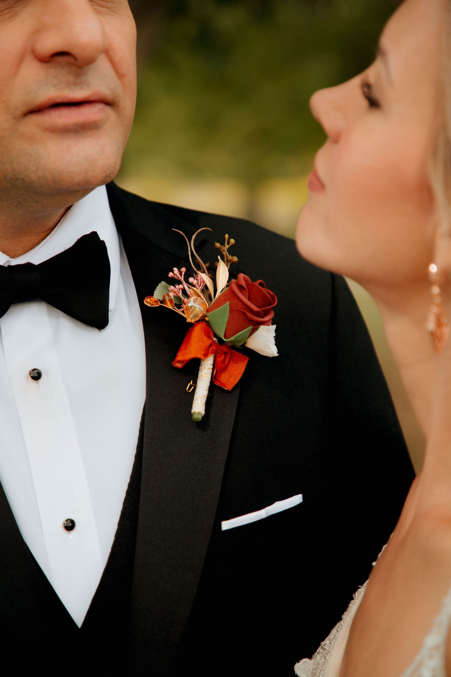 Groom in black tuxedo with fall boutonniere of red rose and orange blooms beside bride.