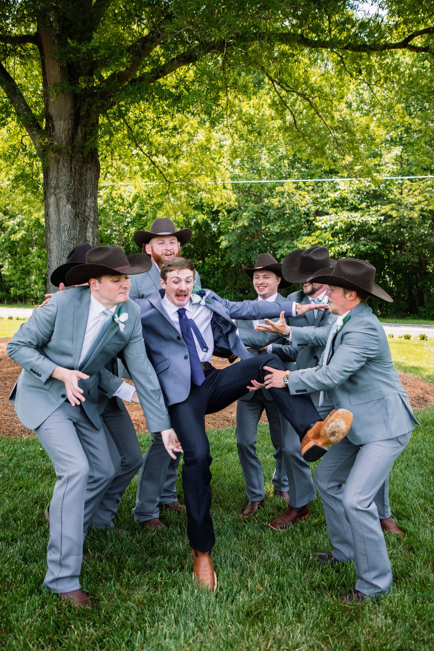 Groomsmen in gray suits and cowboy hats playfully surround the groom in a fun outdoor wedding party photo.