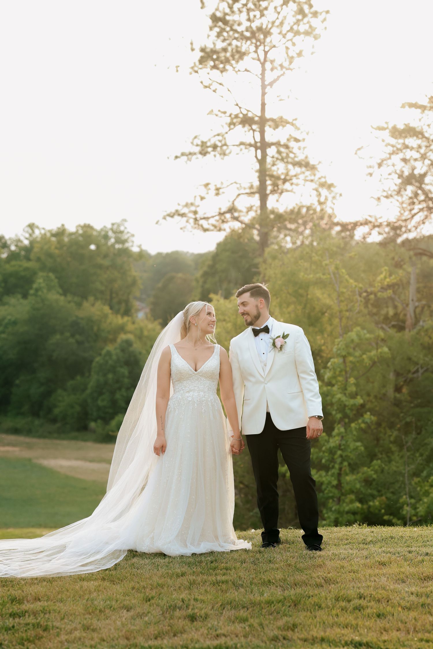 Bride and groom holding hands at golden hour in a lush outdoor wedding setting, groom in white tuxedo jacket.