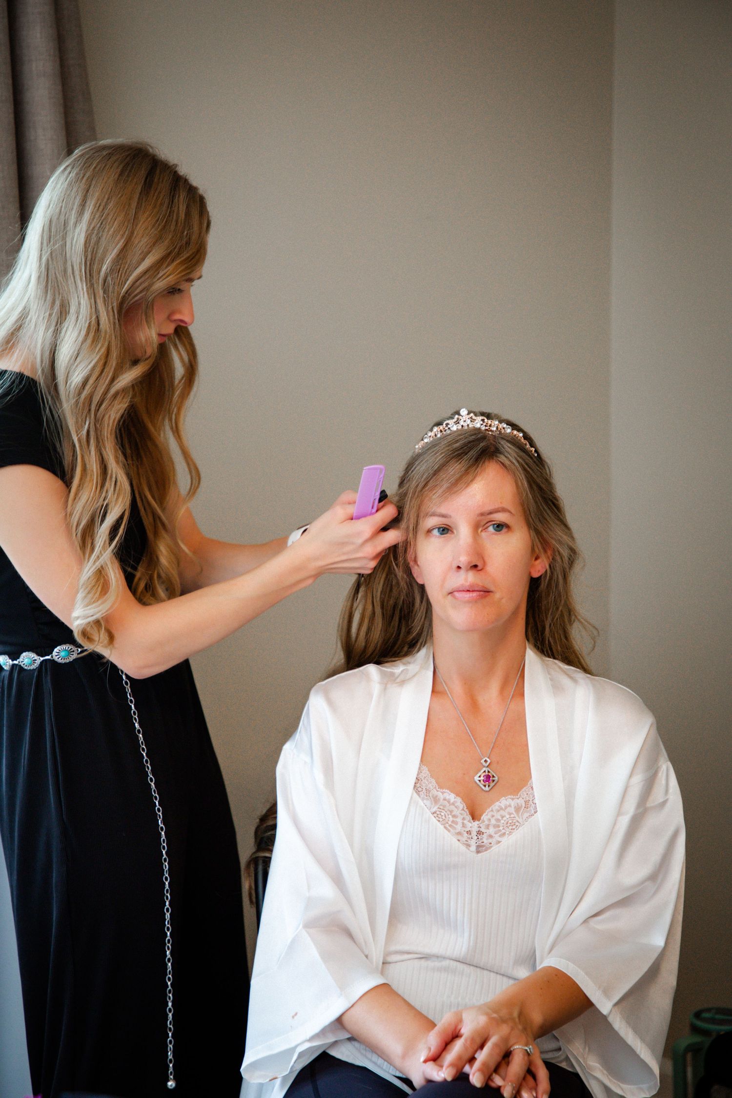 Hairstylist in black dress styling a bride wearing a tiara and white robe.