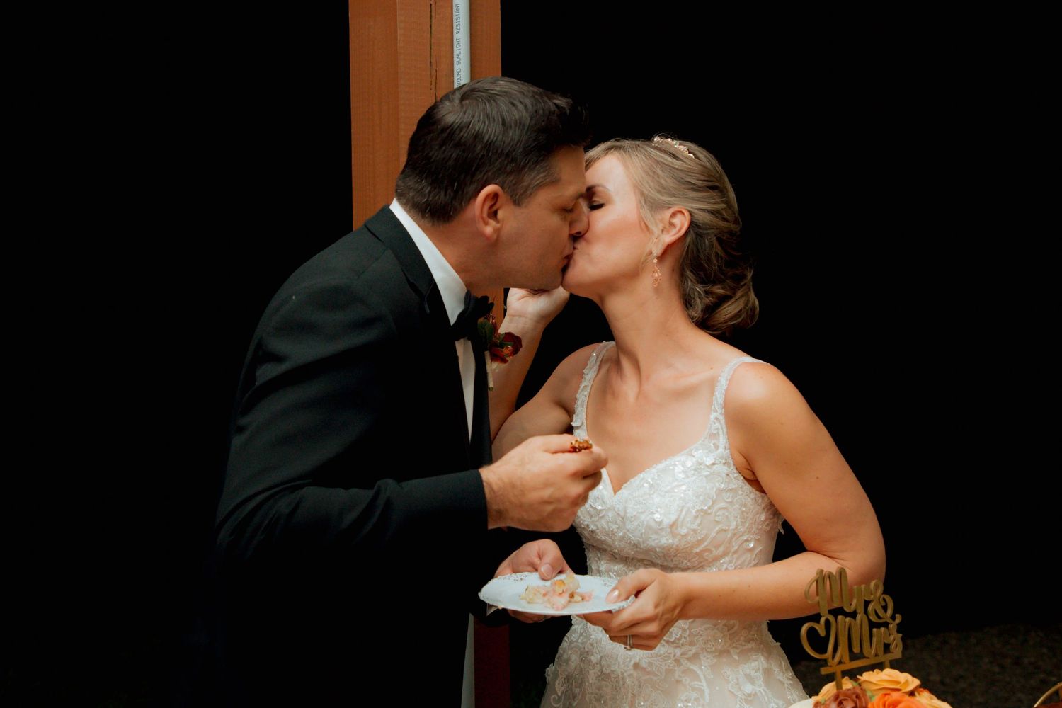 Newlyweds share a kiss while holding cake plates at their wedding reception.