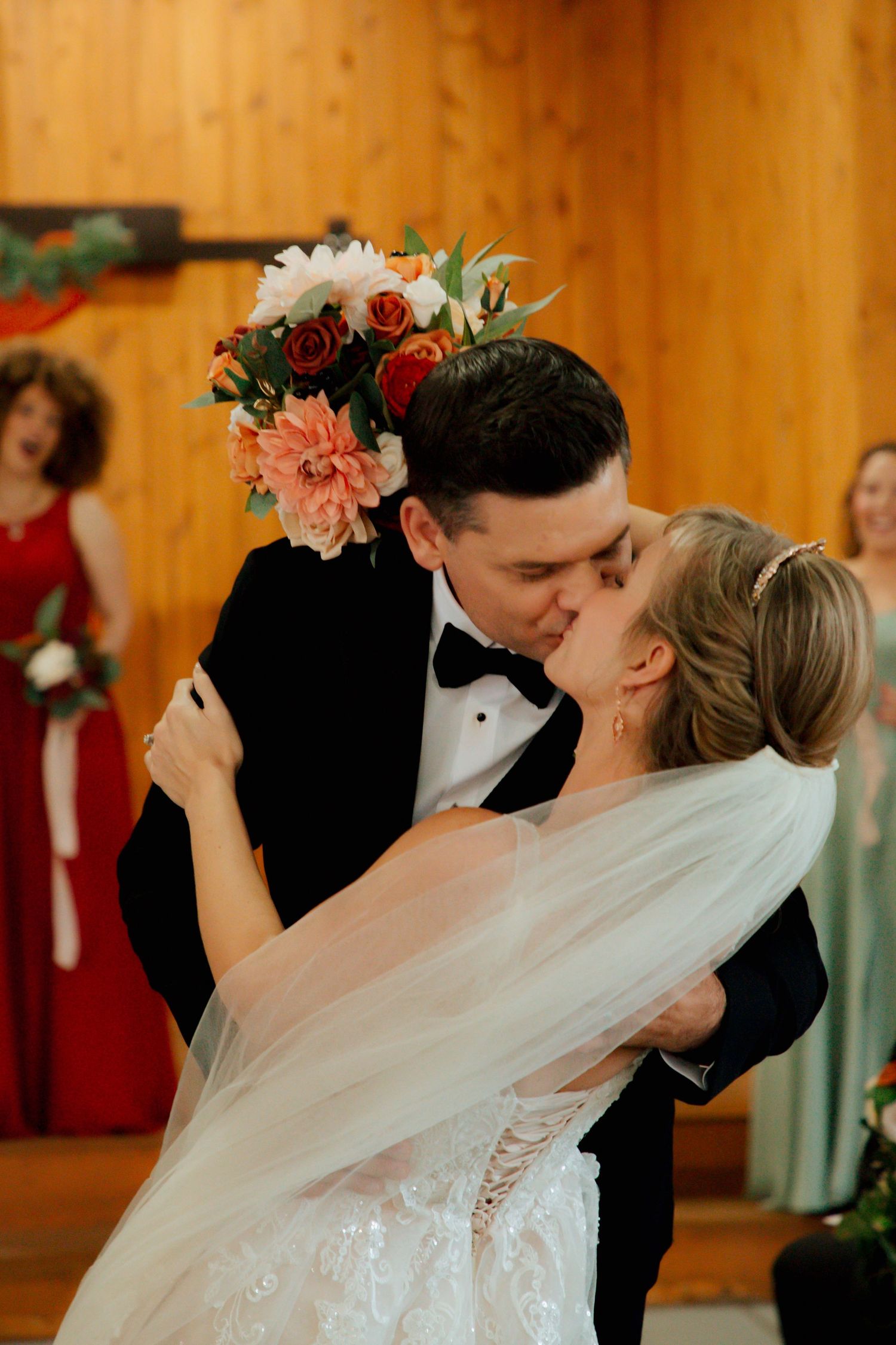 Newlyweds share a passionate kiss as bride holds a vibrant bouquet of coral, red, and white flowers at wedding ceremony.