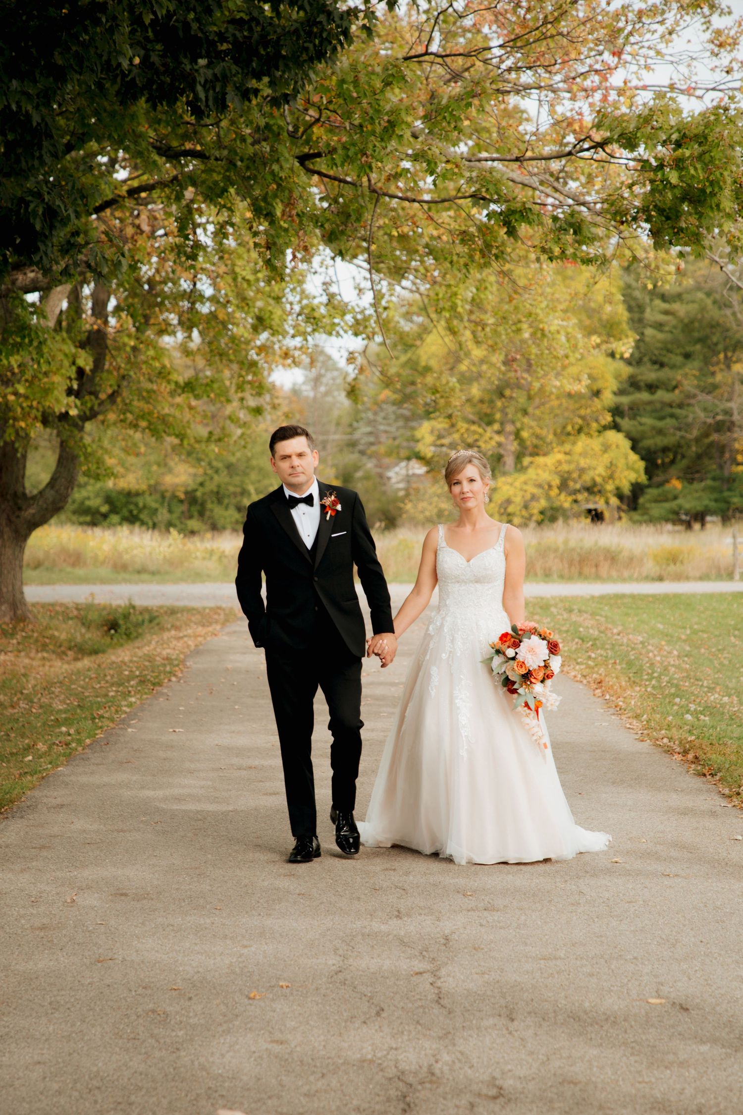 Bride and groom walk hand in hand on autumn path with colorful fall foliage.