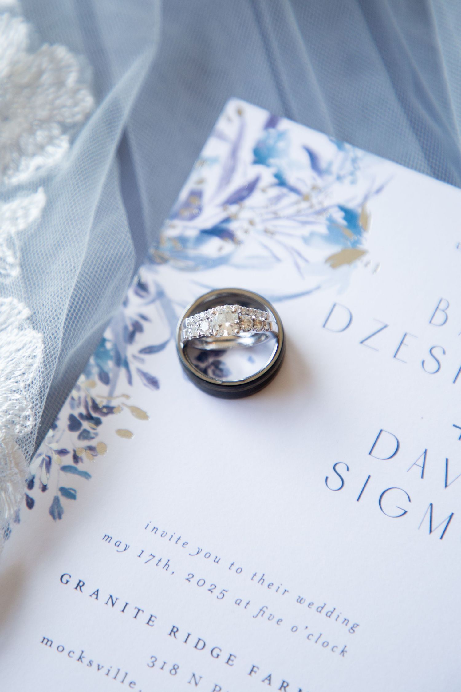Close-up of wedding rings resting on a blue floral wedding invitation for Bailey Dzeskewicz and David Sigmon.