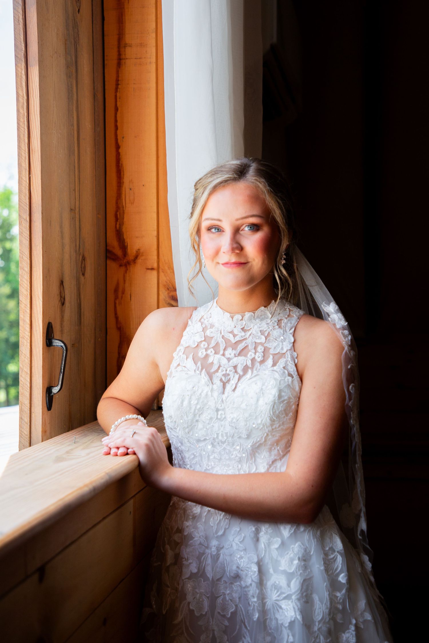 Bride in lace halter wedding gown with veil poses by rustic wooden window in warm natural light.