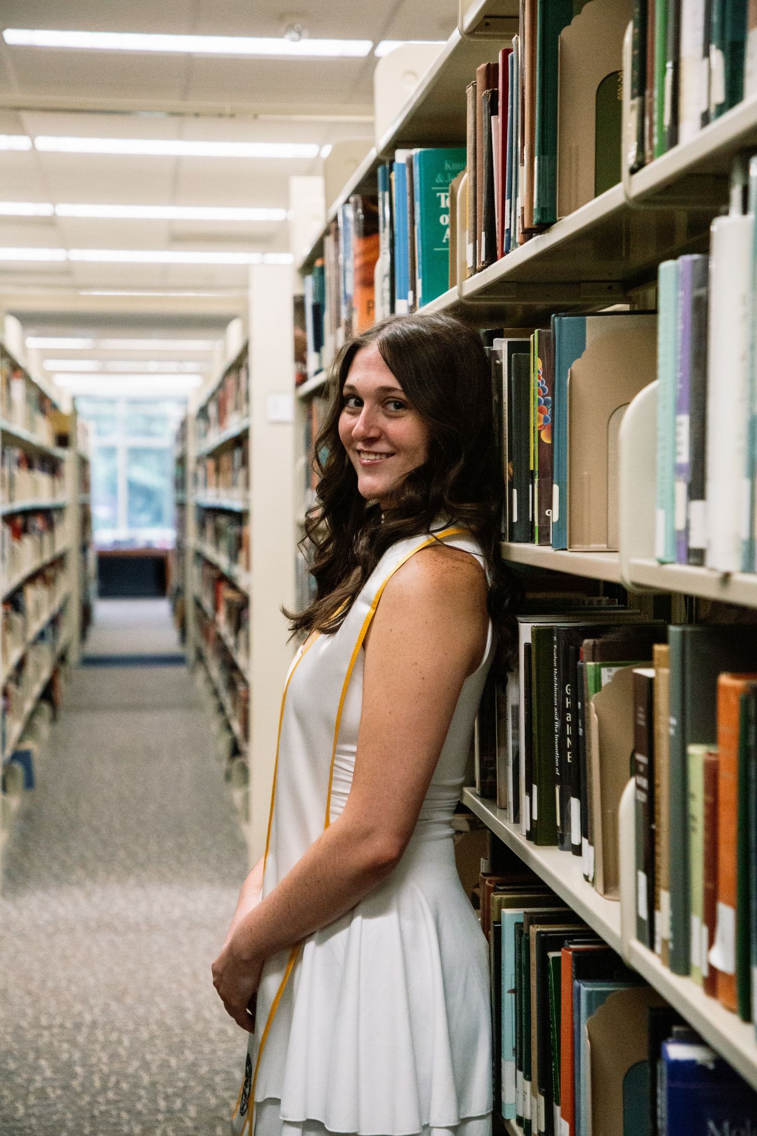 Smiling female graduate in white dress and gold stole stands beside library bookshelf.